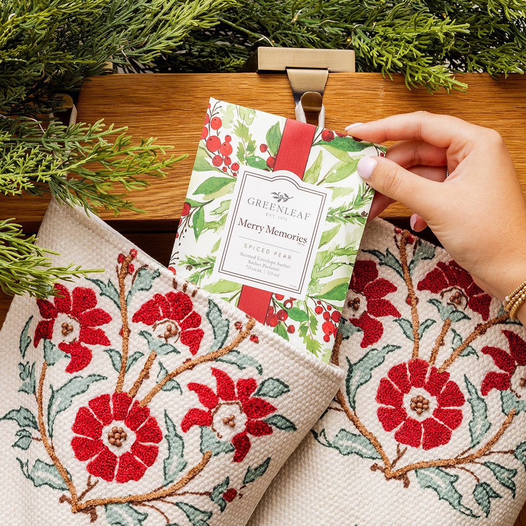 Hand holding a Greenleaf Merry Memories sachet with floral design on a wooden surface with embroidered fabric and greenery. Hand is placing the sachet into a stocking.