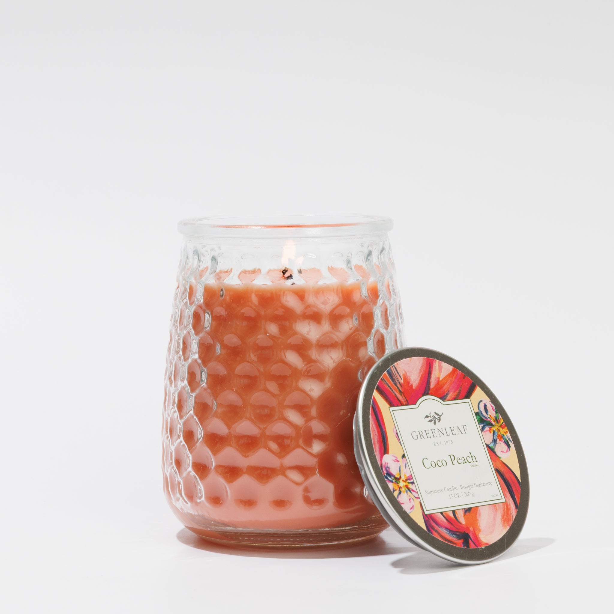 A lit Coco Peach Signature Candle by Greenleaf Gifts sits in a textured pink-orange glass jar with its lid beside it. The floral label is visible against a plain white background.