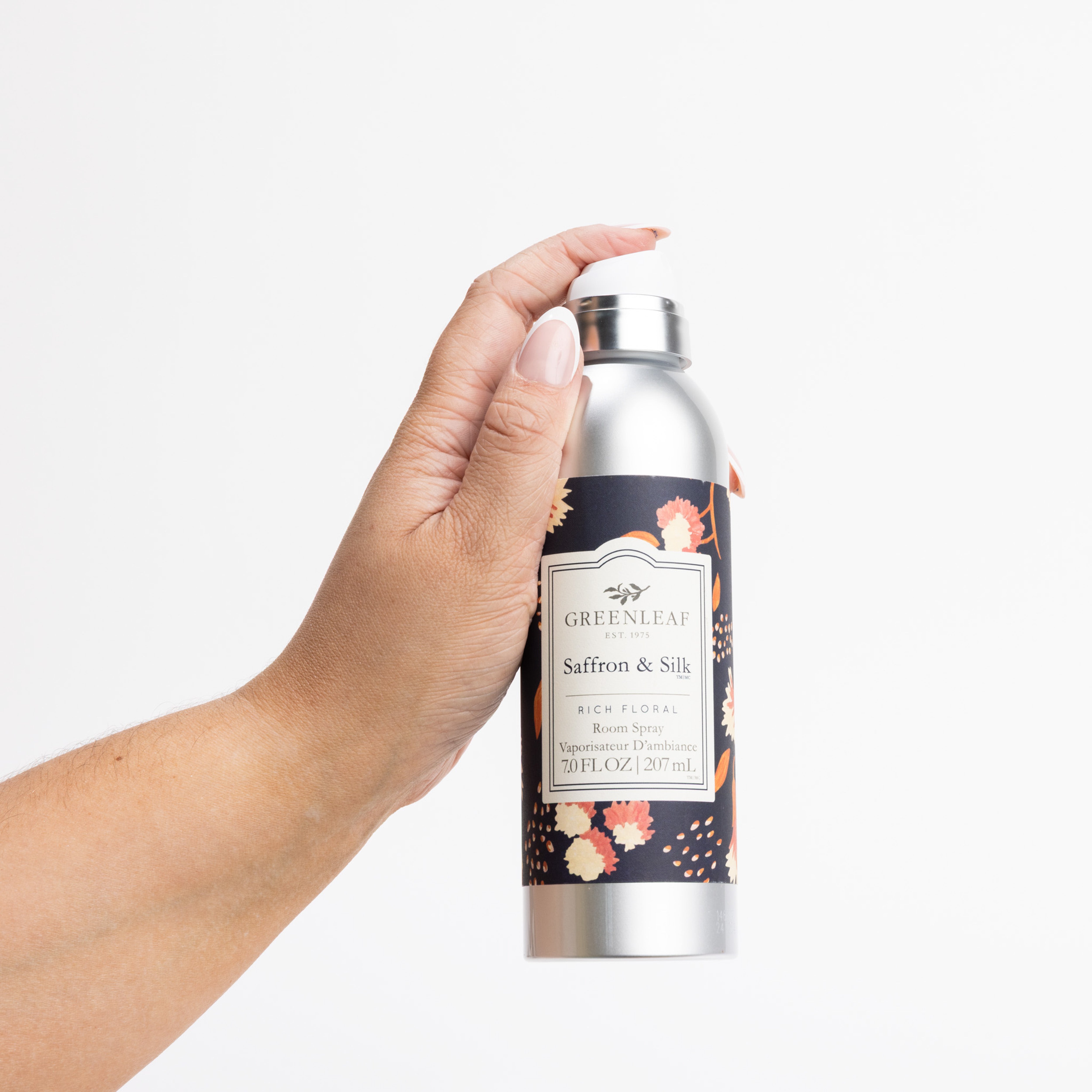 A hand holds a silver bottle labeled Saffron & Silk Room Spray by Greenleaf Gifts, featuring a floral design, against a plain white background. Enjoy the welcoming fragrance of Greenleaf in any room.