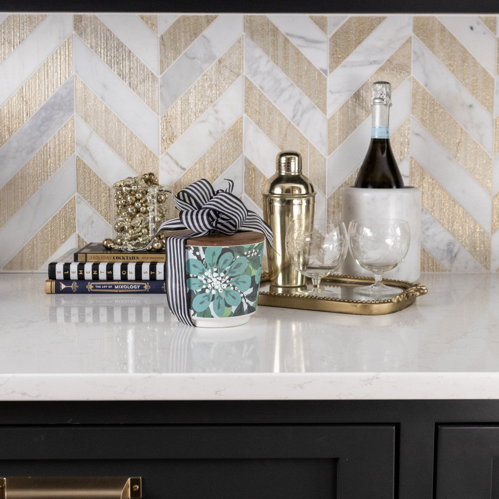 A modern kitchen counter showcases a gold tray with wine, glasses, shaker, books, and the Greenleaf Gifts Shimmering Snowberry Patterned 3 Wick Candle, wrapped in a black and white ribbon, all before a chevron-patterned backsplash.