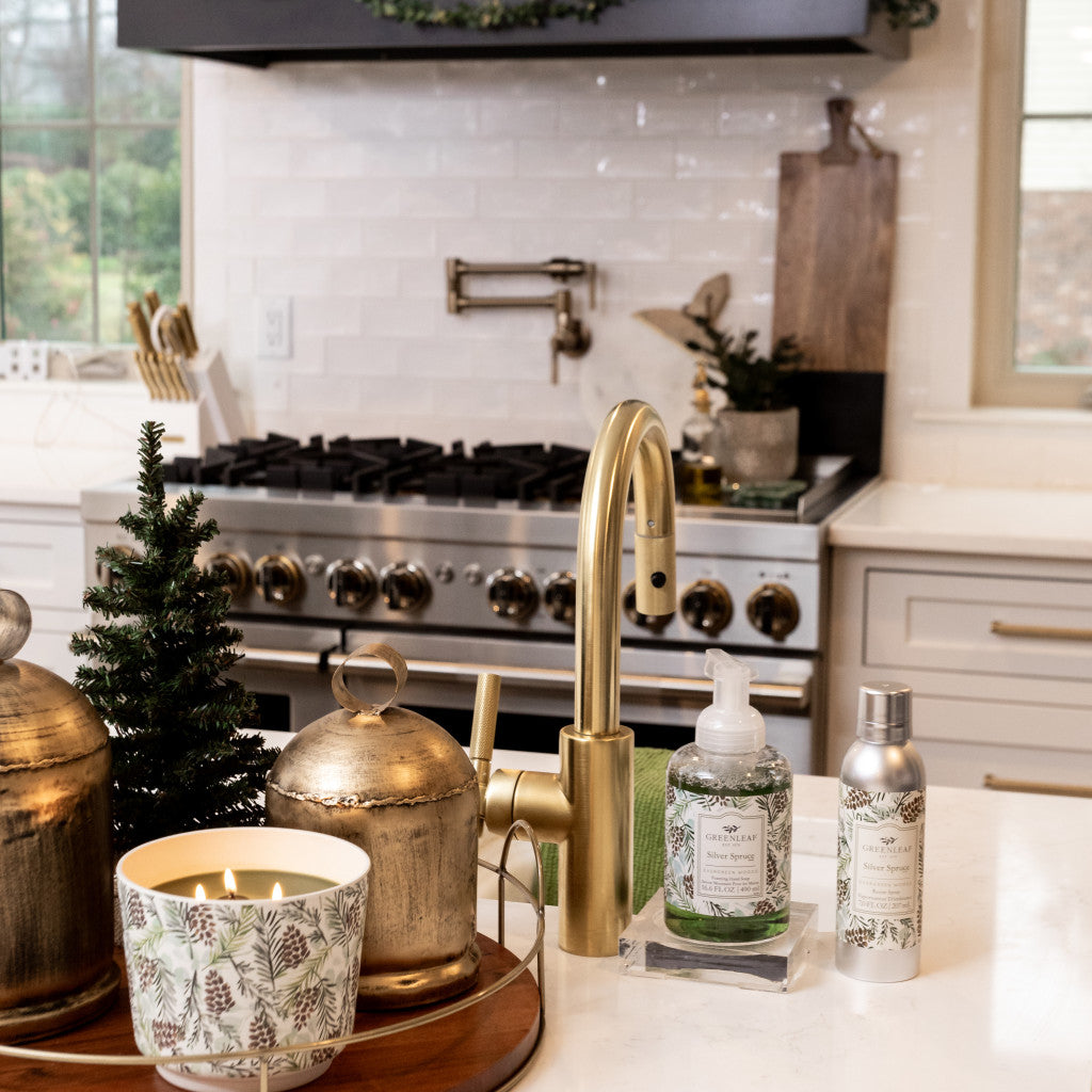 A cozy kitchen features gold fixtures, a small pine tree, and the Greenleaf Gifts Silver Spruce Patterned 3 Wick Candle. Botanical-labeled soap and lotion bottles rest on the white countertop beside other patterned candles.