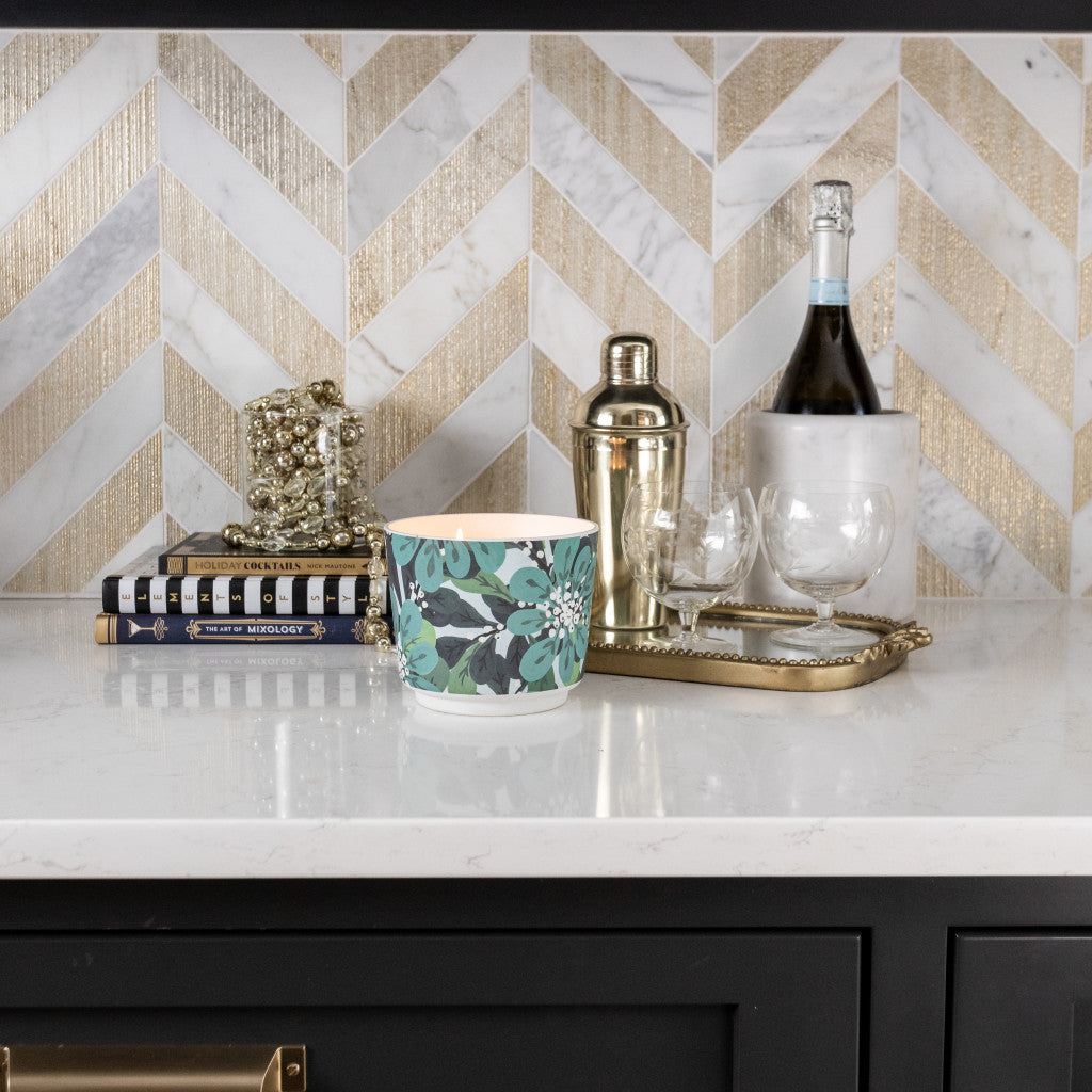 A marble countertop displays the Greenleaf Gifts Shimmering Snowberry Patterned 3 Wick Candle, stacked books, jewelry, a gold cocktail shaker and tray, two glasses, sparkling wine, all set before a gold and white chevron tile backsplash.