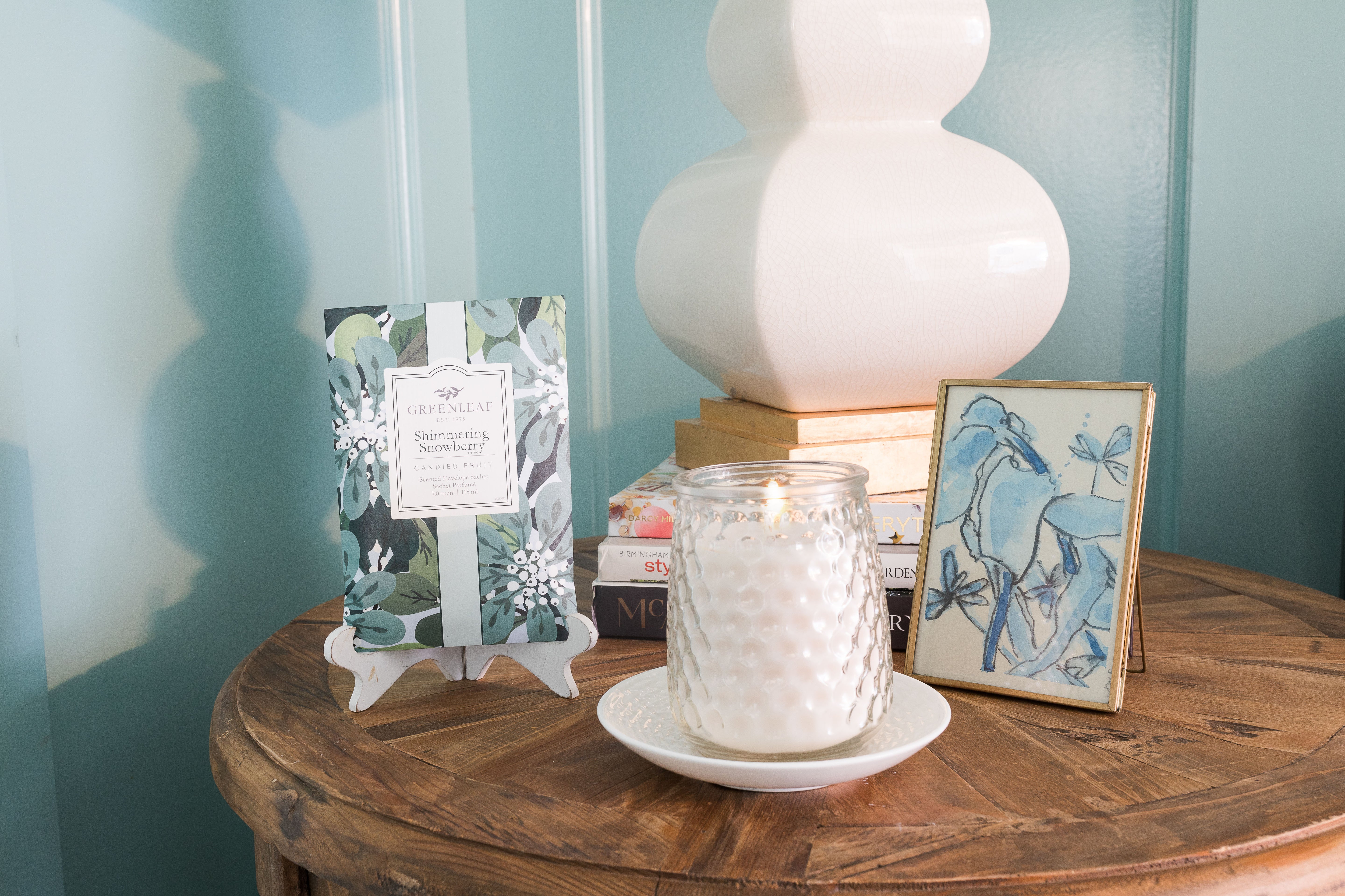 A round wooden table holds a Greenleaf Gifts Shimmering Snowberry Signature Candle in a glass jar on a saucer, a packet of matching sachet, stacked books, a lamp, and a framed blue floral print set against a teal wall.