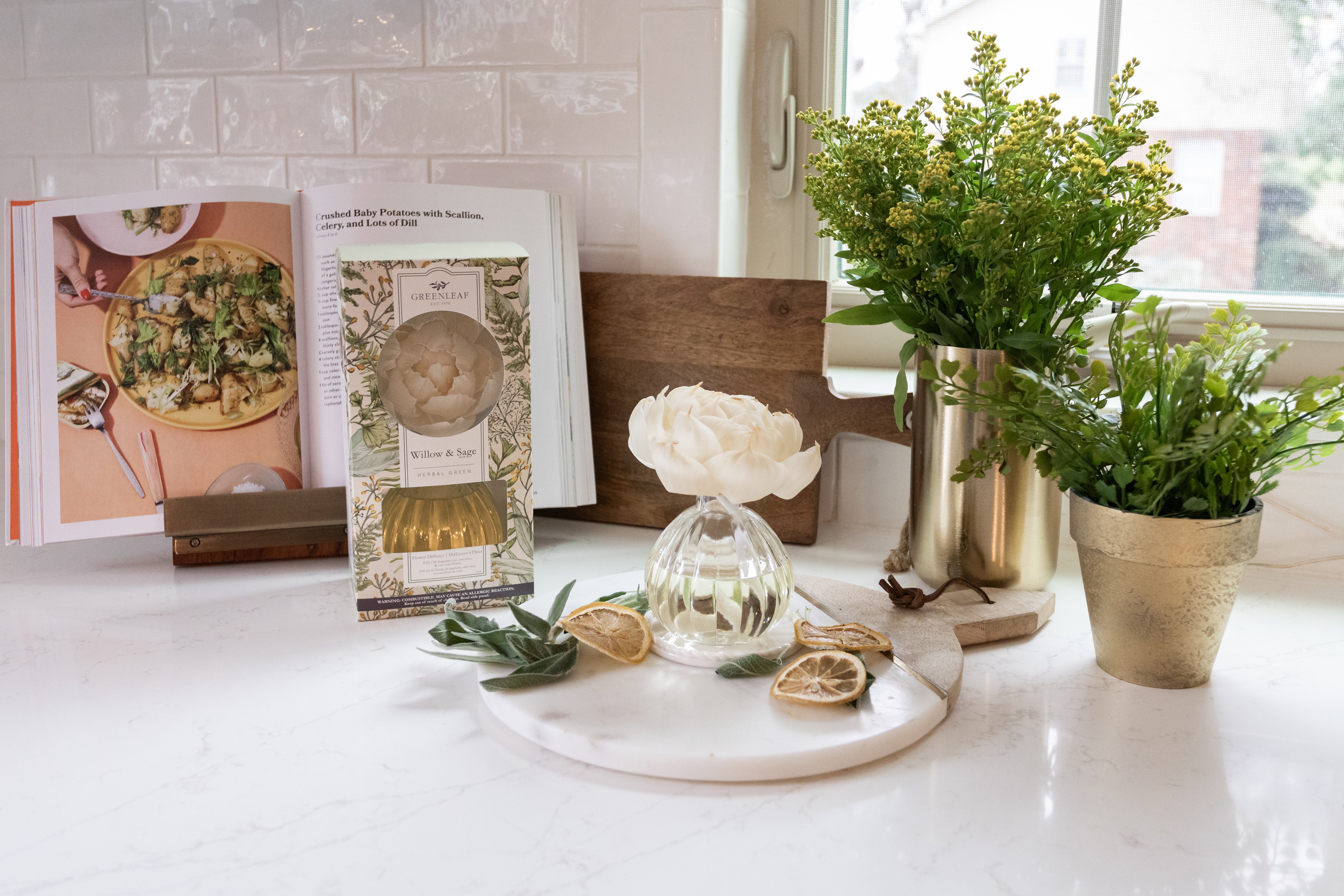 A bright kitchen counter displays an open cookbook, a Greenleaf Gifts Willow & Sage Flower Diffuser, a small vase with a white flower, dried citrus slices, a gold pitcher, and potted greenery on a marble tray.