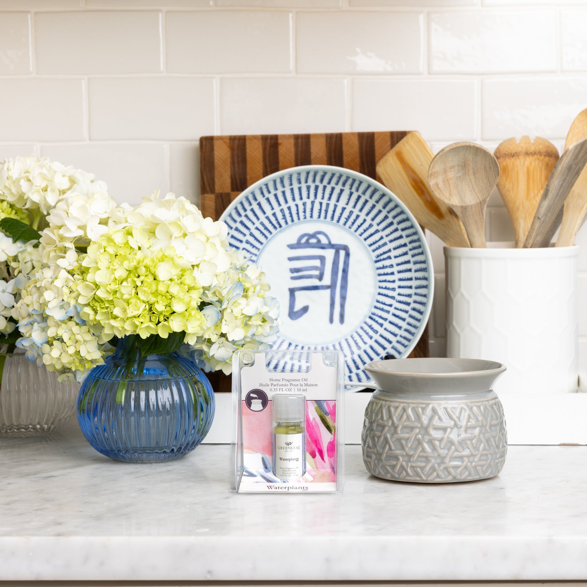 A kitchen counter displays a vase of white and green hydrangeas, a blue patterned plate, wooden utensils in a white holder, a gray ceramic warmer for flameless fragrance, and Greenleaf Gifts Waterplants Home Fragrance Oil on marble.