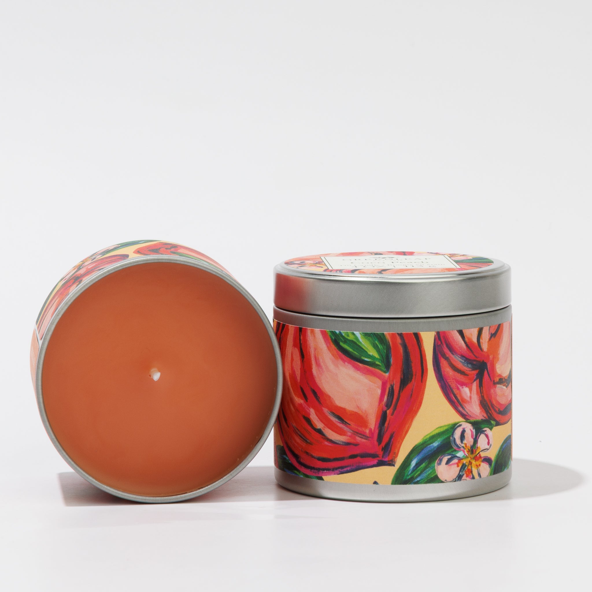 Two decorative Greenleaf Gifts Coco Peach Tin Candles with colorful floral designs: one stands upright with the lid, the other lies on its side, showing unlit orange wax. Both are set against a white background.