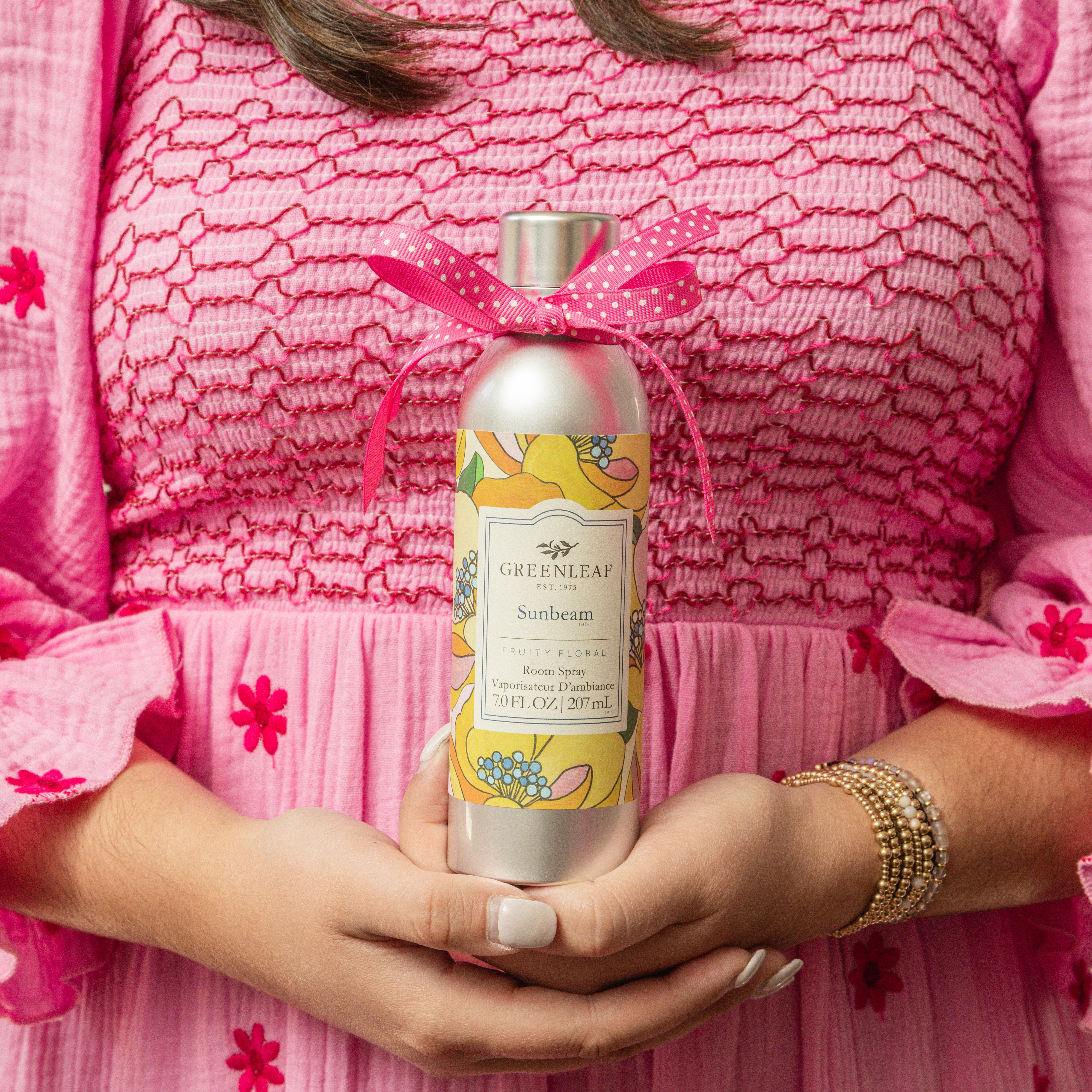 A woman in a pink dress holds a Greenleaf Gifts Sunbeam Room Spray, decorated with a pink ribbon and floral label. Her fingernails are painted pink, and she wears a pearl bracelet.