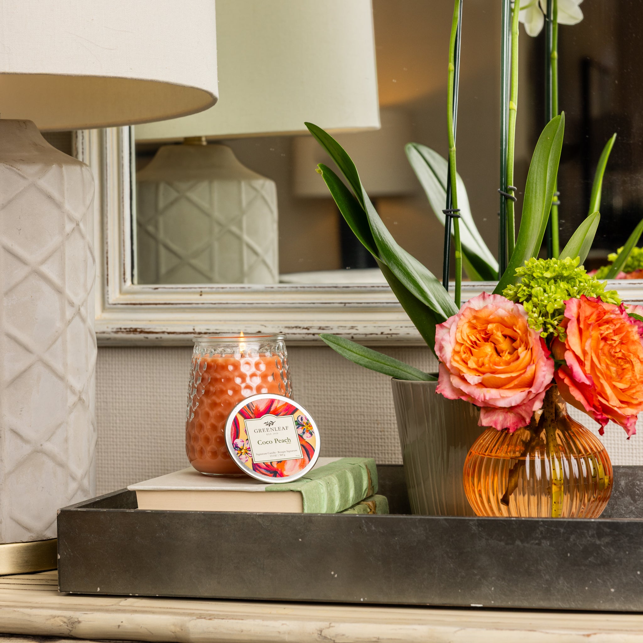 A decorative tray features a lit Greenleaf Gifts Coco Peach Signature Candle, a round Greenleaf tin, stacked books, and an orange ribbed vase with bright flowers and leaves next to a white lamp and mirror.