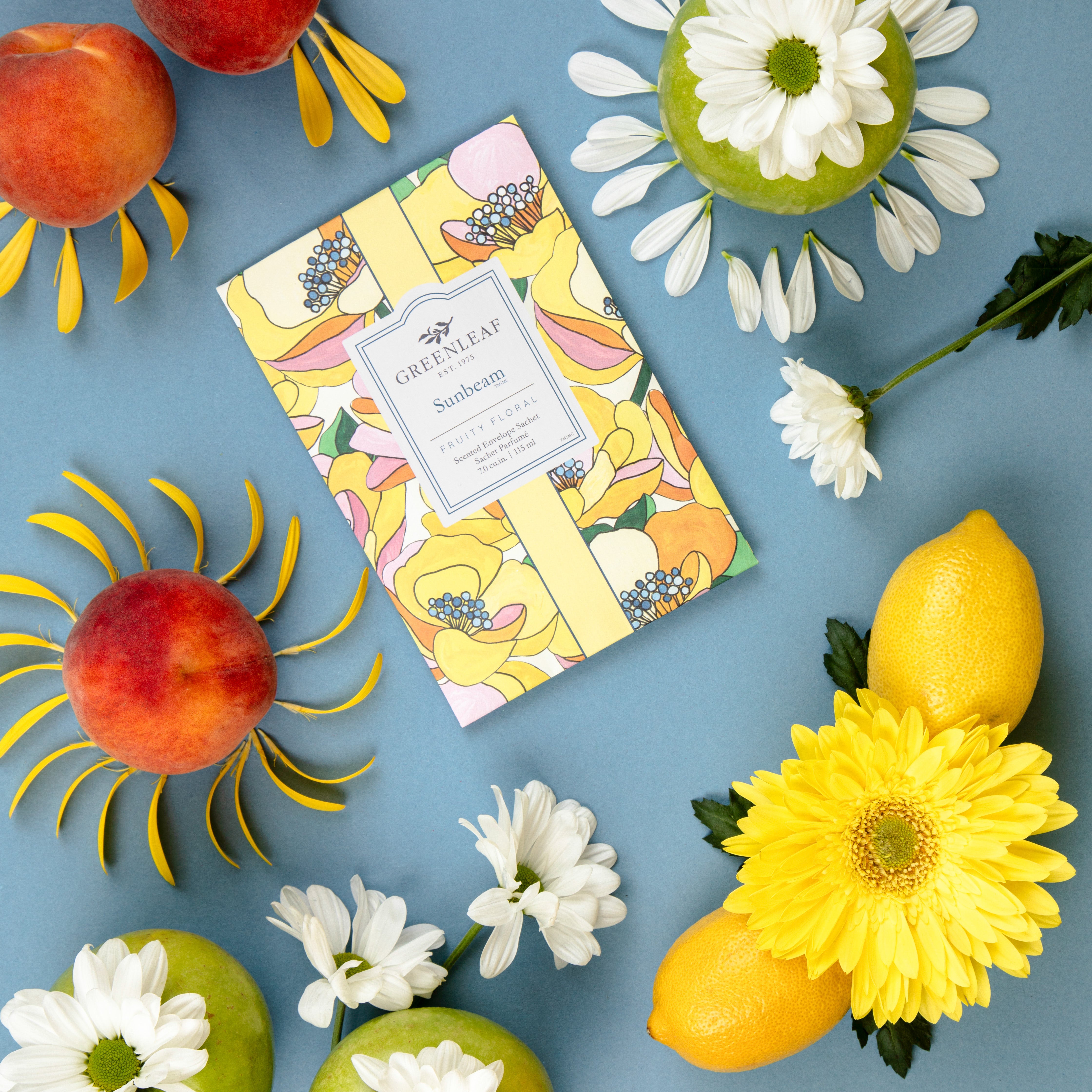 A vibrant flat lay features the Greenleaf Gifts Sunbeam Sachet among peaches, lemons, and yellow daisies on a blue background. Petal-adorned fruits create a fresh vibe, highlighting this long-lasting air freshener.