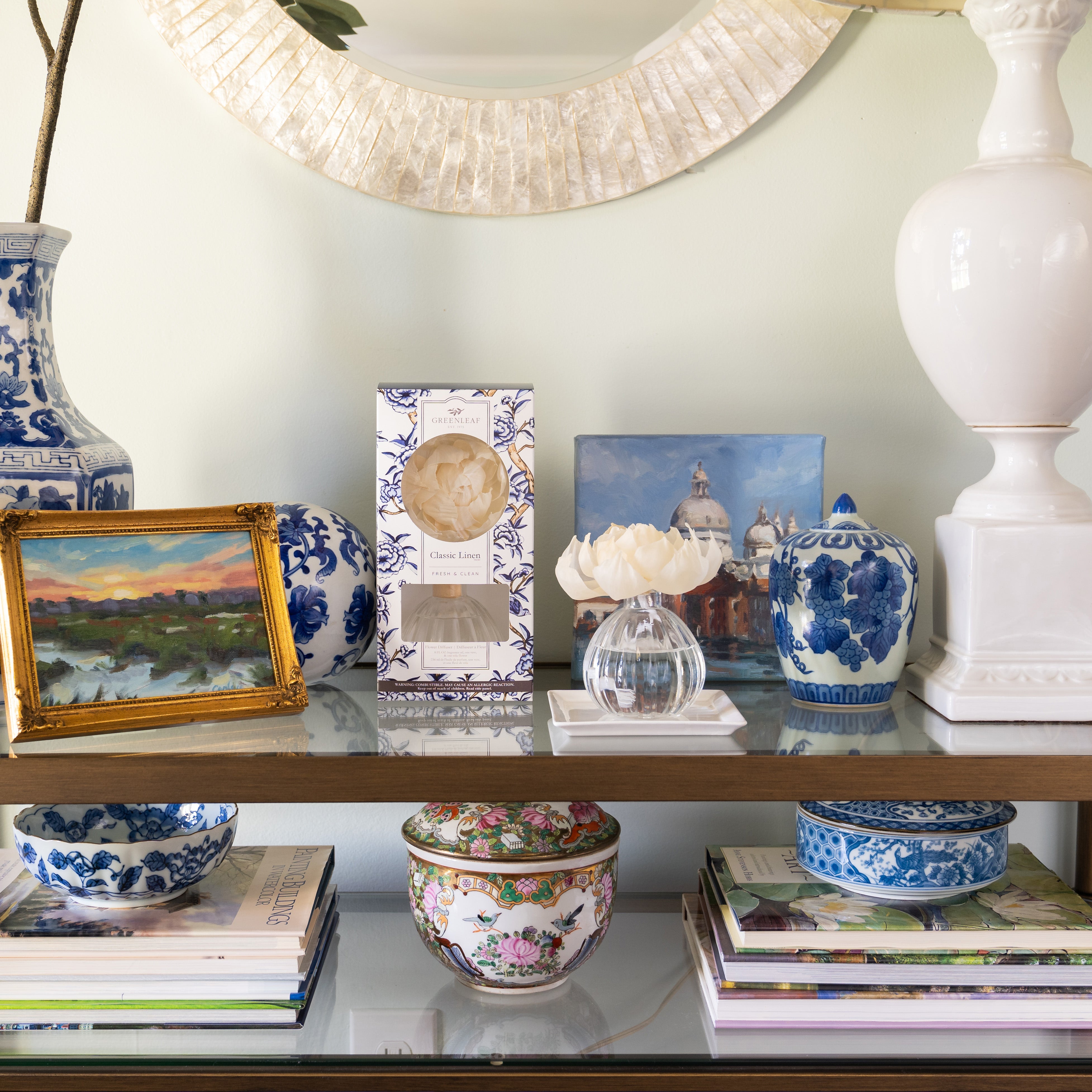A shelf displays blue-and-white ceramics, small landscape paintings, books, a white vase, a Greenleaf Gifts Flower Diffuser-Classic Linen (GLG966498S) with a white flower, and a decorative box—arranged elegantly for 60 days of freshness.