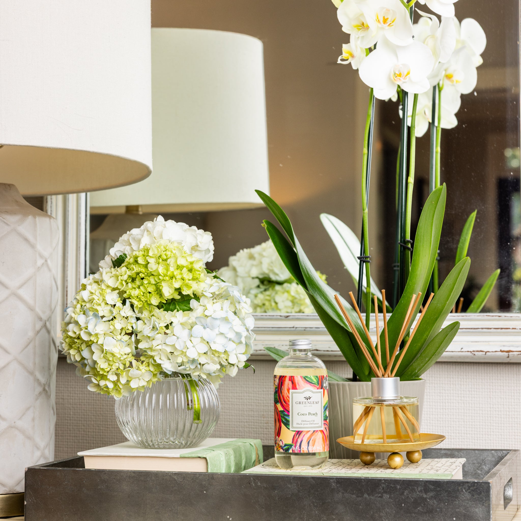 A table features a glass vase with white and green hydrangeas, a Greenleaf Gifts Coco Peach Diffuser Oil, a candle labeled “Citrus Peach,” a book, and a white lamp. A mirror and white orchids are in the background.