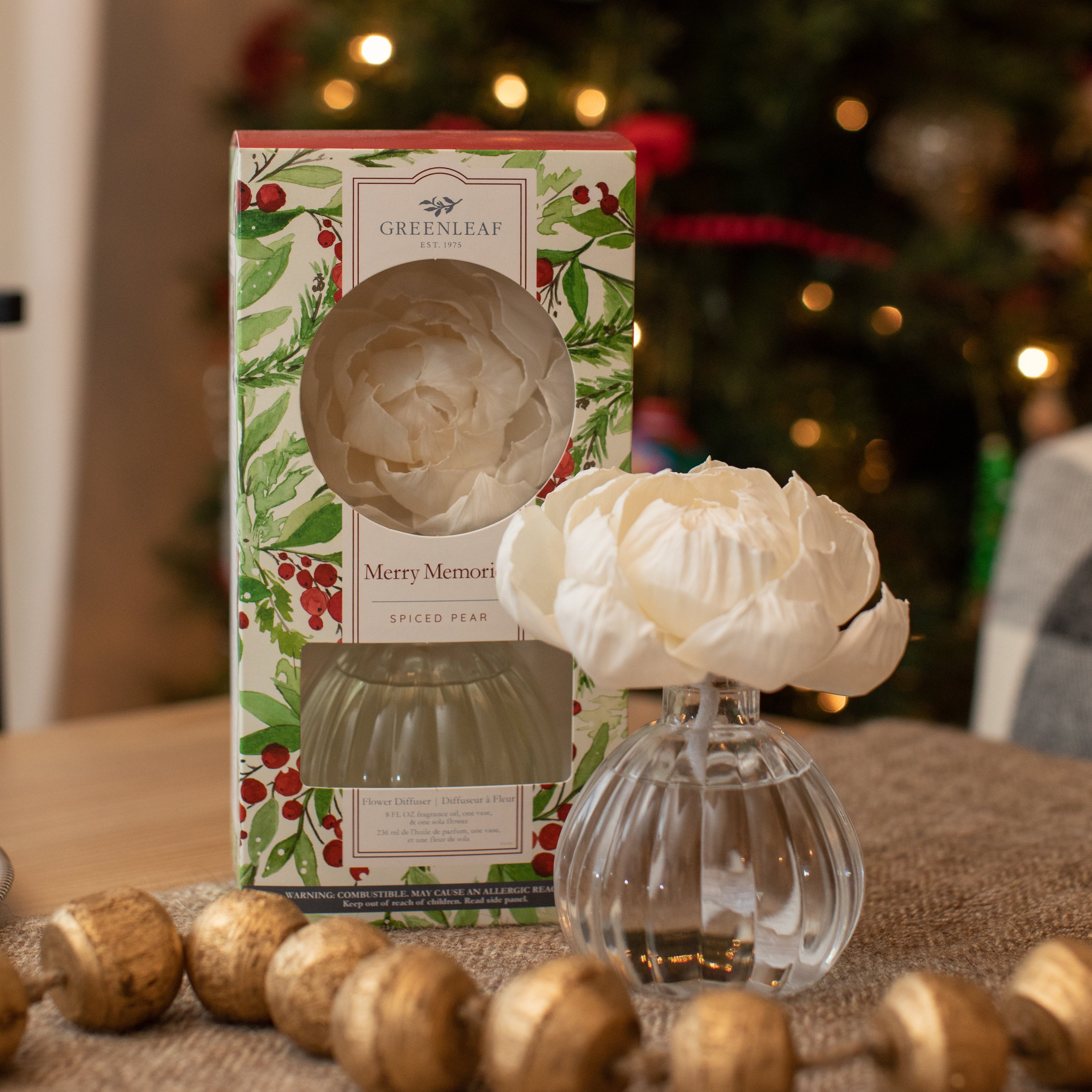 A boxed 8 oz Greenleaf Gifts Merry Memories Flower Diffuser sits beside an open glass diffuser with a white flower, surrounded by holiday decor and a softly glowing Christmas tree. Fragrance lasts up to 60 days.