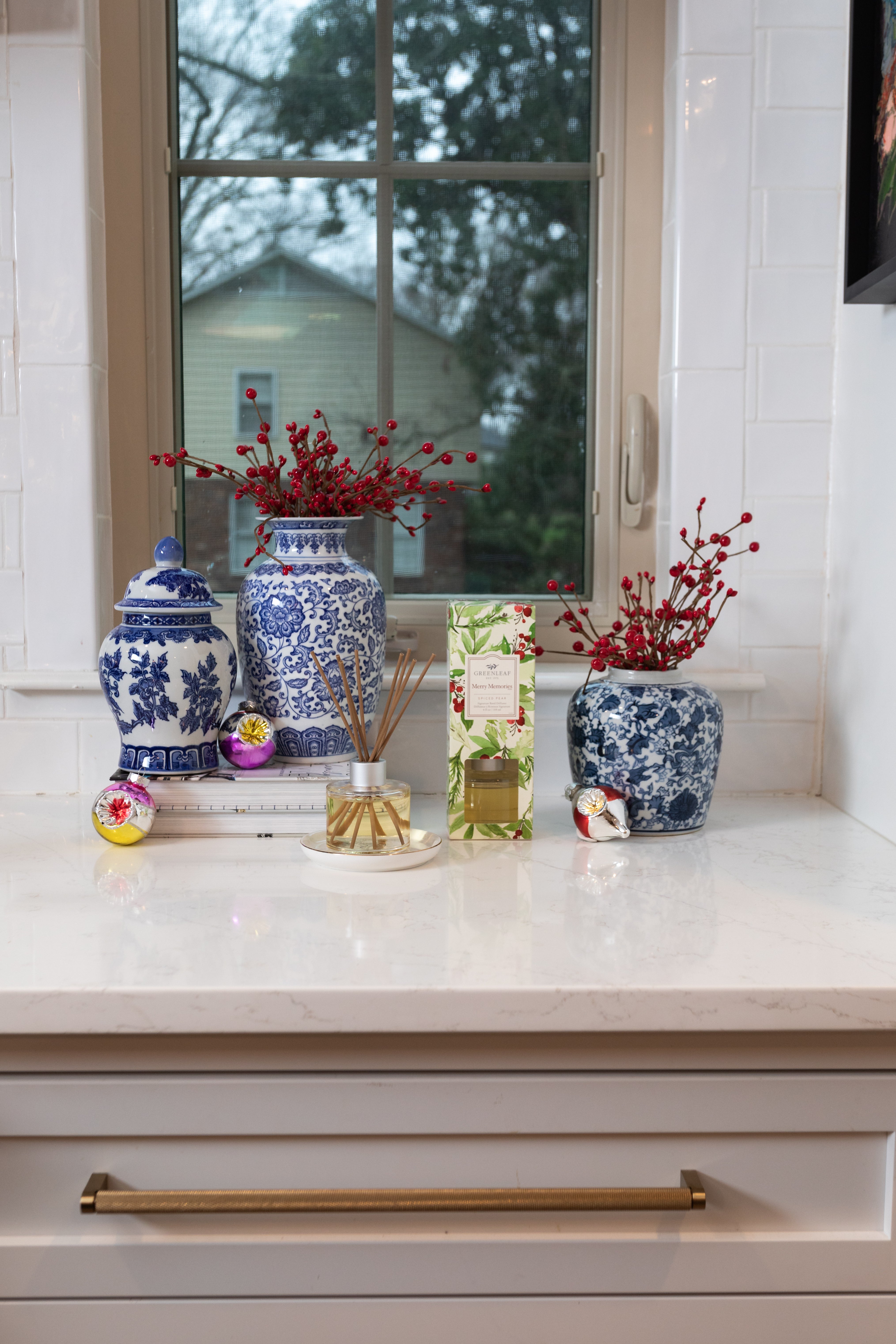 Blue and white vases with red berry branches sit on a marble countertop below a window. Also on the counter are festive ornaments and the Silver Spruce Reed Diffuser by Greenleaf Gifts with fiber reeds.
