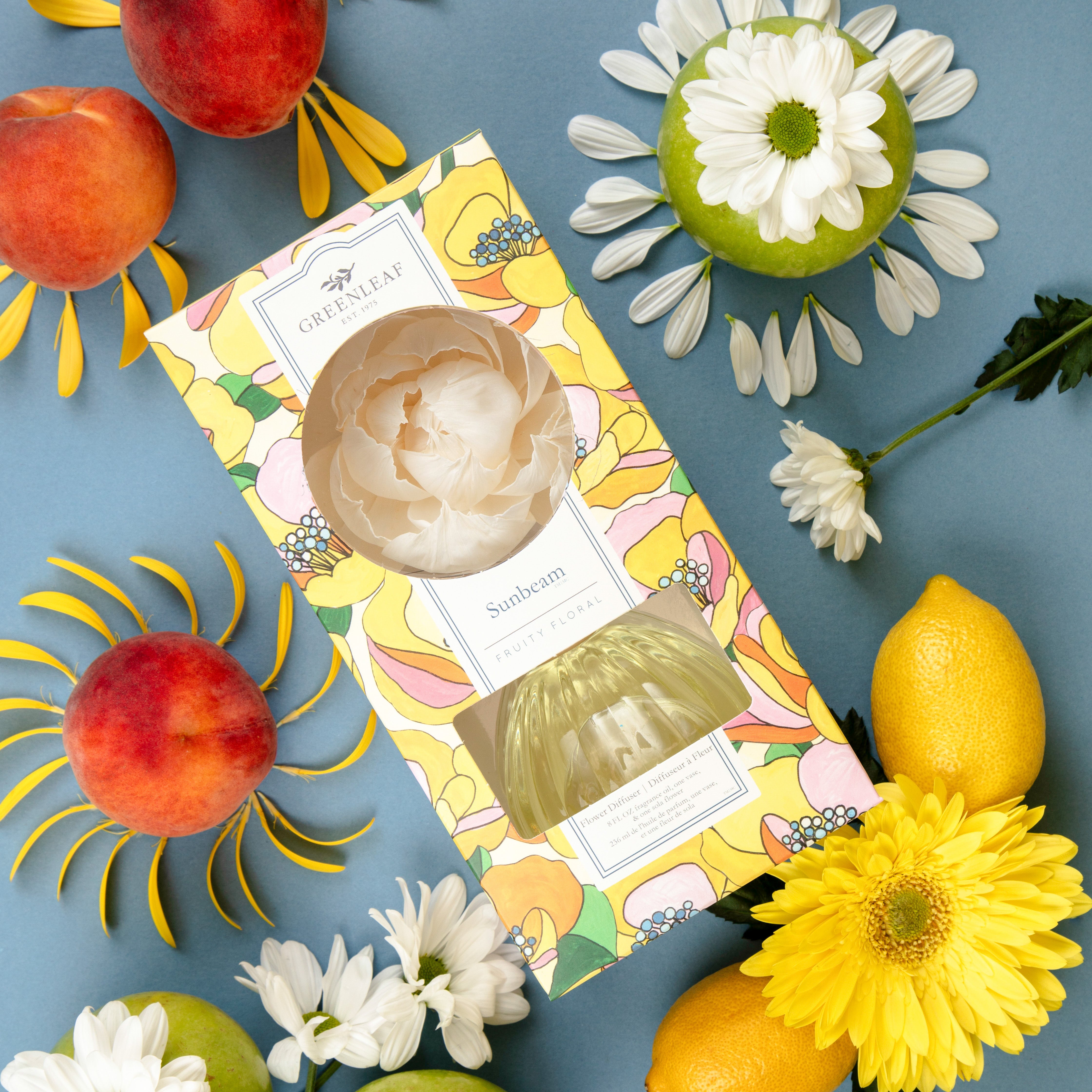 A vibrant flat lay featuring a Greenleaf Gifts Sunbeam Flower Diffuser, peaches, lemons, daisies, yellow flowers, and petals arranged in sunburst patterns on a blue background.
