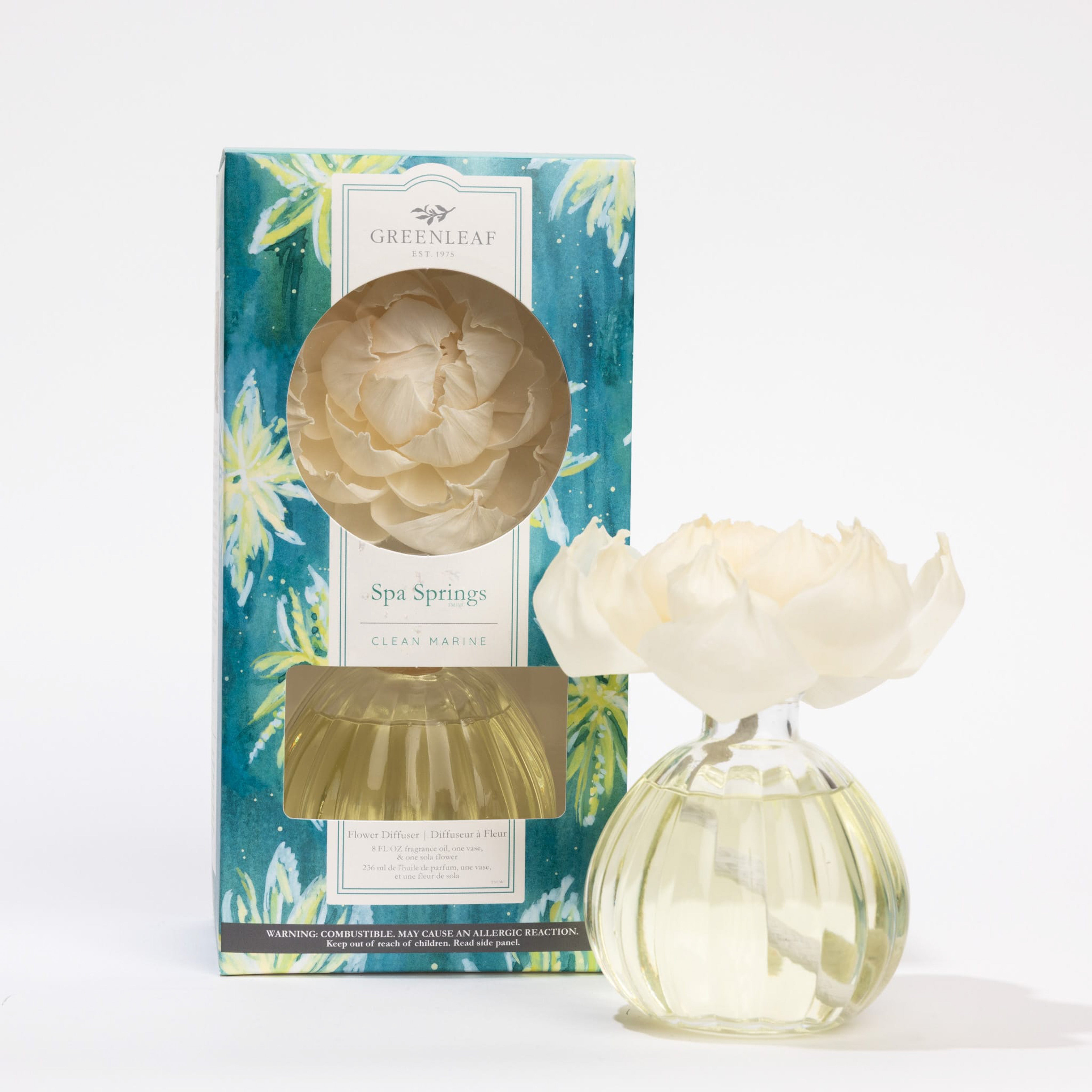 A Greenleaf Gifts Spa Springs Flower Diffuser, 8 oz, features a decorative white flower on a round glass bottle and lasts up to 60 days. It is displayed beside its floral-patterned box.