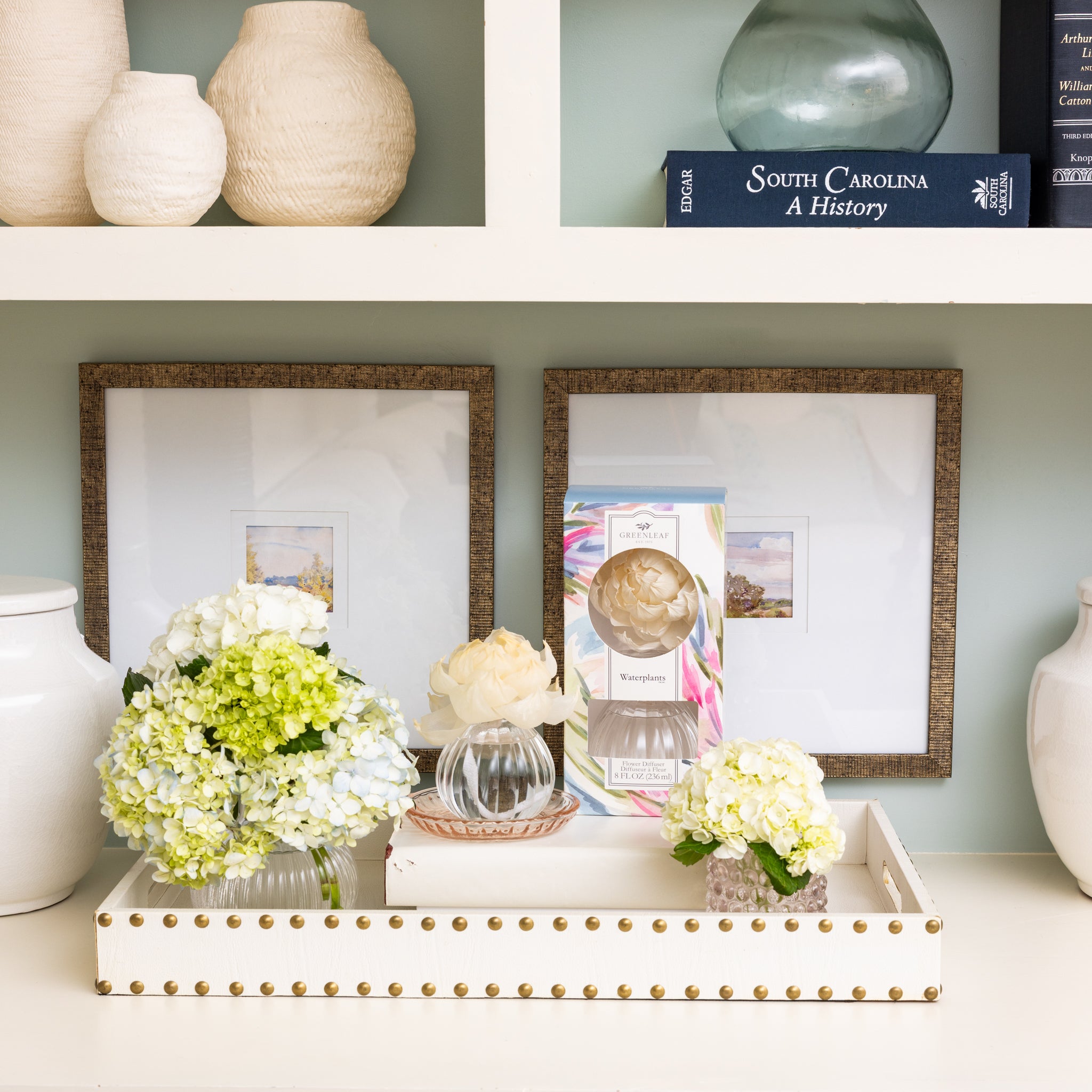 A neat, light-filled decorative shelf display features framed art, white vases, a book, glass bowls with artificial flowers, and the Greenleaf Gifts Waterplants Flower Diffuser for a fresh touch.