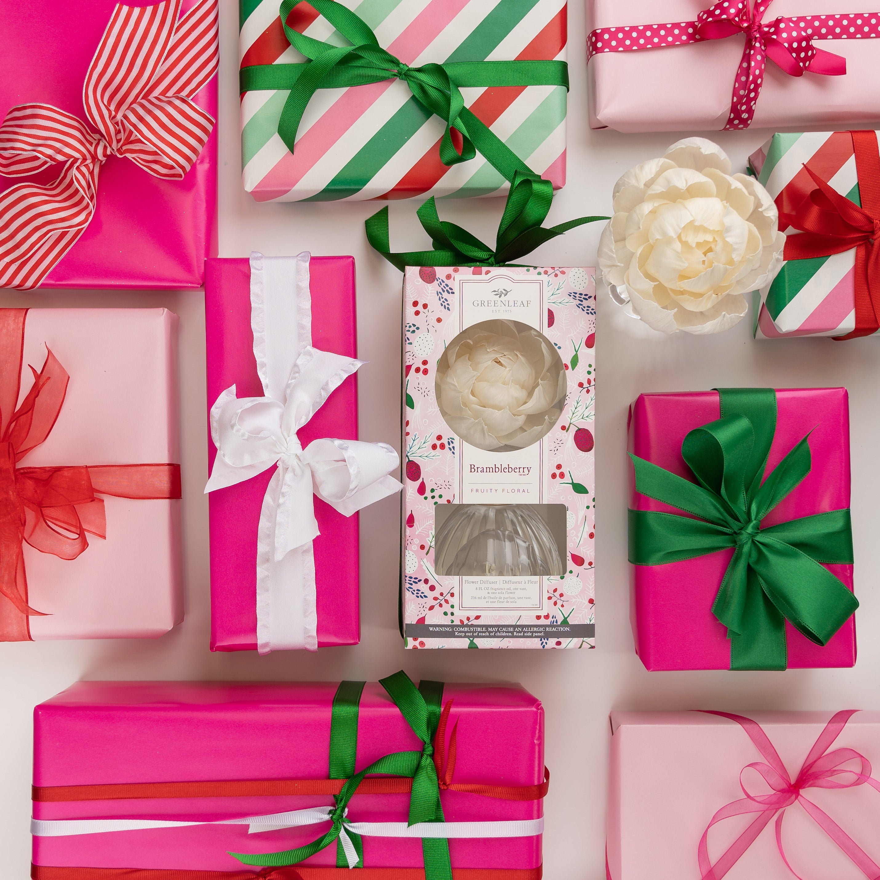An array of pink, red, and green wrapped gift boxes with ribbons surrounds a Greenleaf Gifts Brambleberry Flower Diffuser and a white flower decoration. The gifts are neatly arranged on a white background.