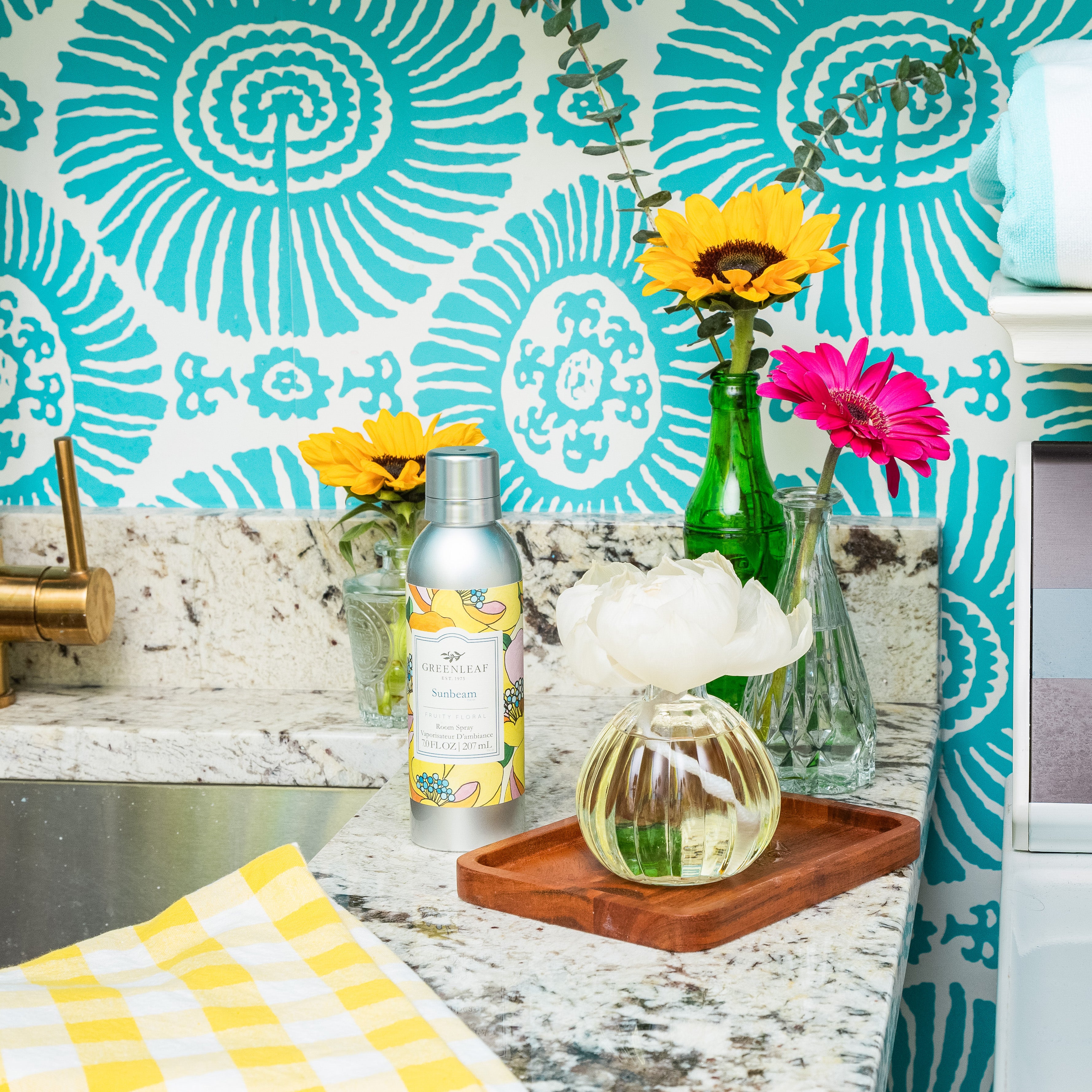 A granite kitchen counter displays a silver Sunbeam Room Spray by Greenleaf Gifts, a small vase with a white flower, yellow and pink flowers in glass vases, a yellow checkered cloth, all set against a turquoise patterned wall.