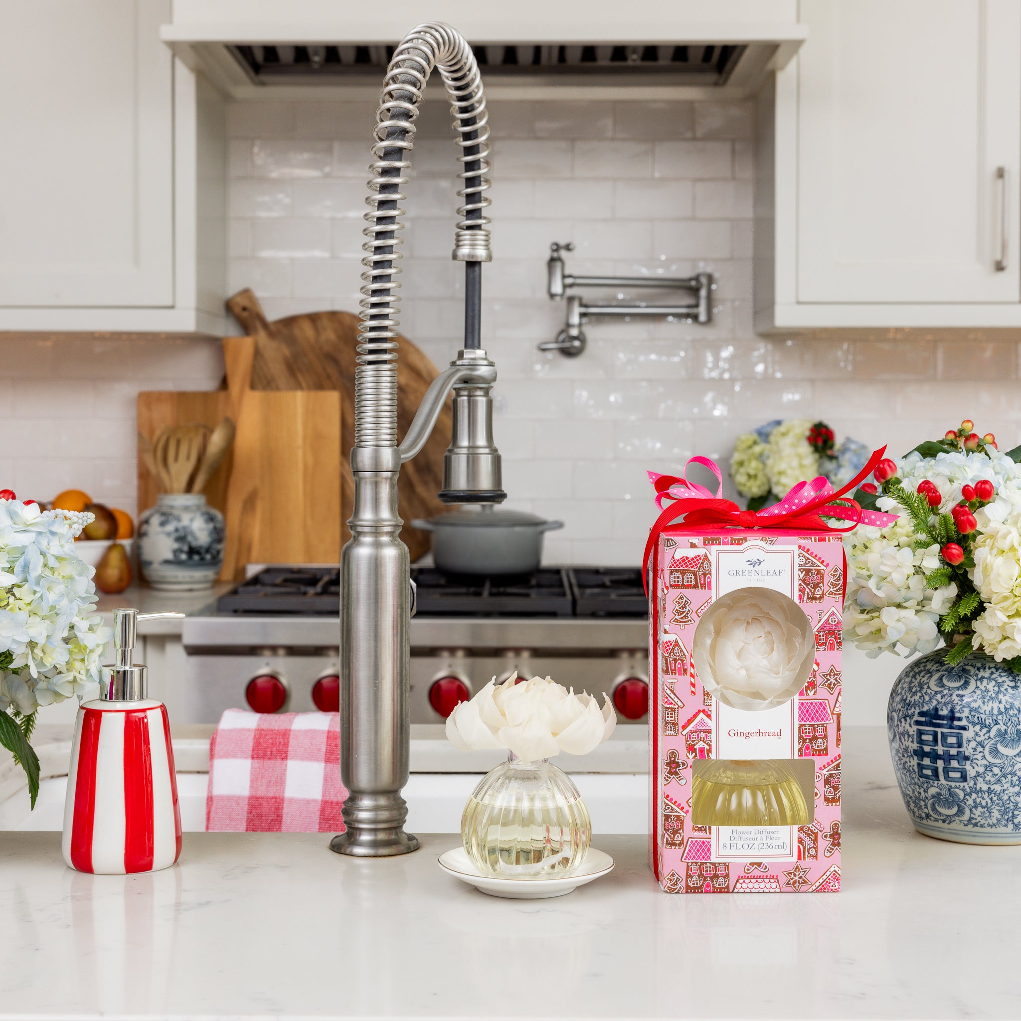 A modern kitchen features a stainless steel faucet, white countertop, flowers in vases, a red and white soap dispenser, and the Greenleaf Gifts Gingerbread Flower Diffuser gift set with sola paper flower near the sink.