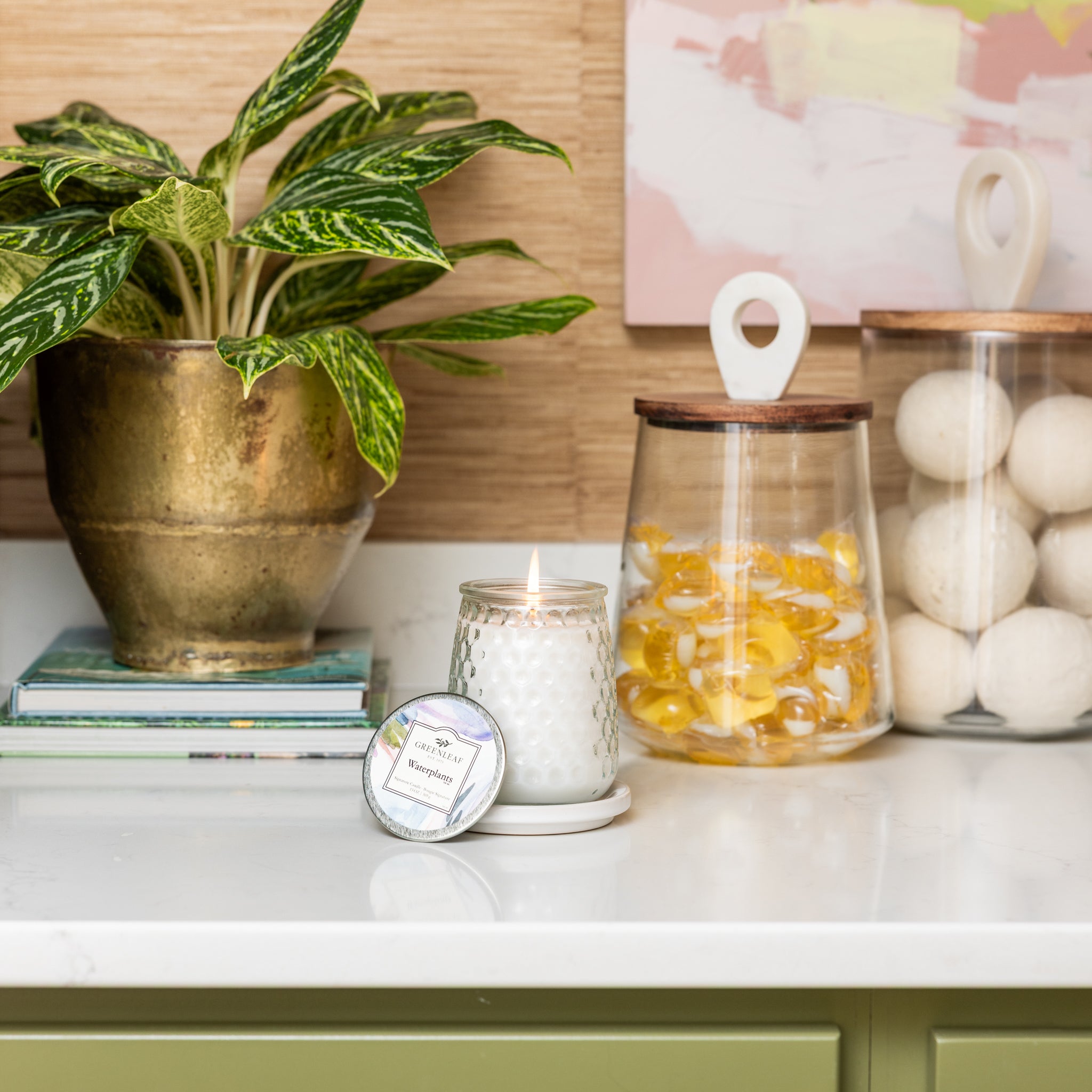 A lit Waterplants Signature Candle by Greenleaf Gifts in a textured glass jar sits on a white countertop beside its lid, near a potted plant, books, and jars with yellow and white decor, infusing the scene with Greenleaf fragrance.