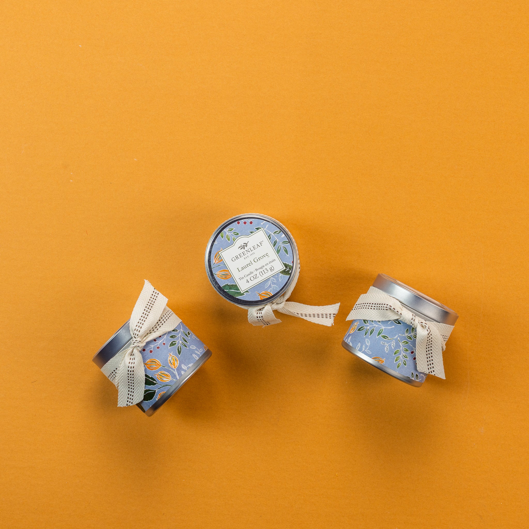 Three small, decorative blue tins with white ribbon bows are arranged on an orange background. Reminiscent of elegant travel candles, the tins feature delicate floral patterns, and one displays a label reading "Laurel Grove Tin Candle, Greenleaf Gifts, 113g / 4oz"—a delightful nod to Greenleaf fragrance.