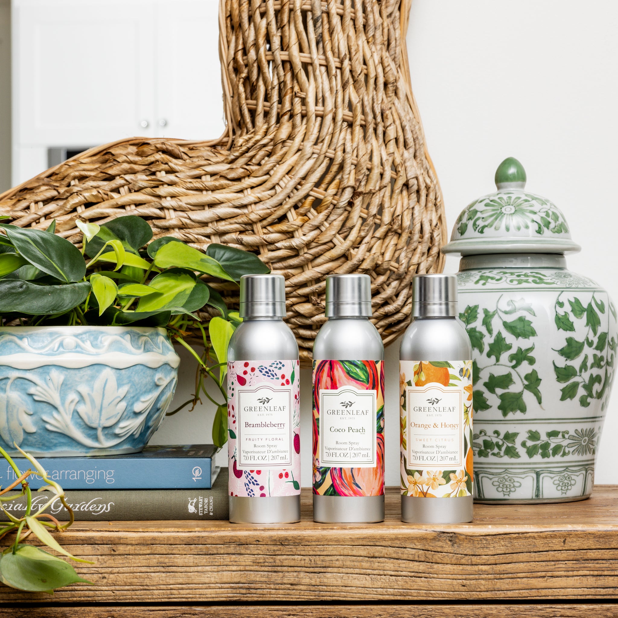 Three metallic spray bottles, including the Greenleaf Gifts Coco Peach Room Spray, are displayed on a wooden table beside a blue planter with green leaves, a wicker accent, stacked books, and a green-and-white ceramic jar.