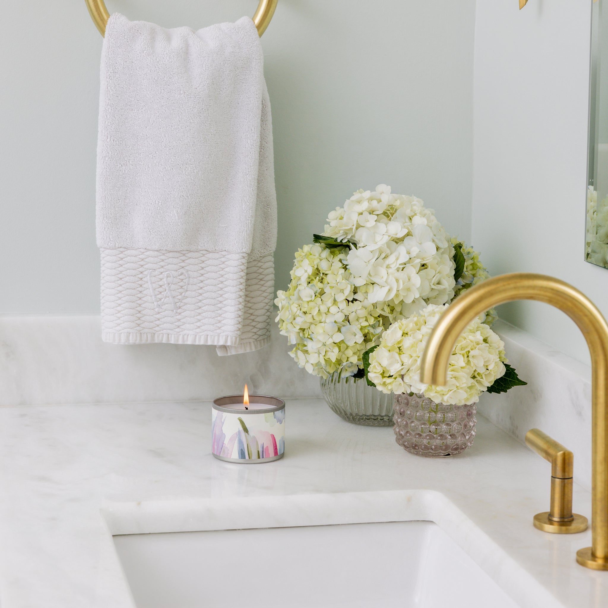 A white towel hangs on a gold ring above a marble bathroom counter featuring the Waterplants Tin Candle by Greenleaf Gifts, a vase of white hydrangeas, and a gold faucet by the sink.