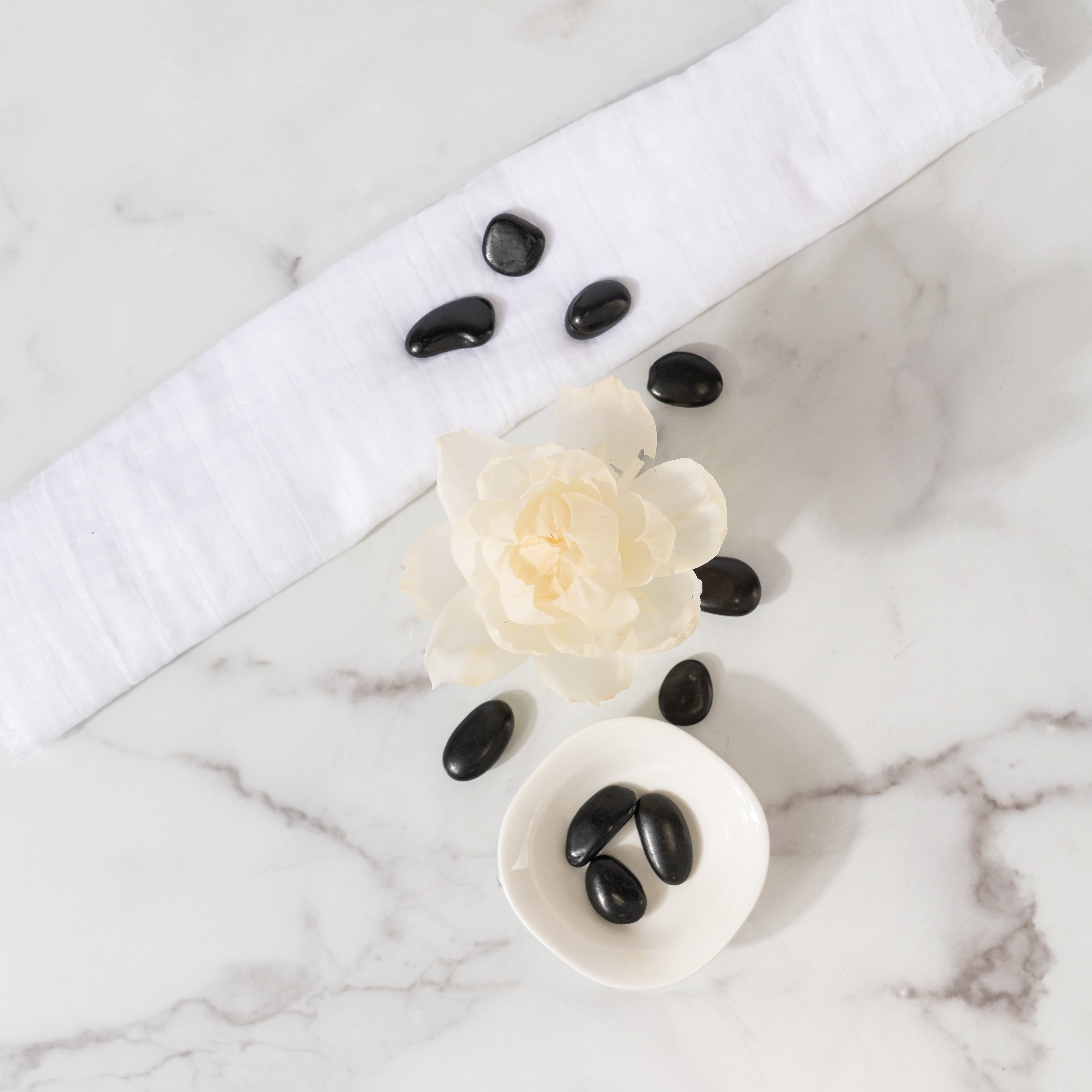 A white marble surface displays Greenleaf Gifts' Spa Springs Flower Diffuser (8 oz) with a folded towel, cream flower, smooth black stones, and a small dish of stones—perfectly styled for a serene setting.