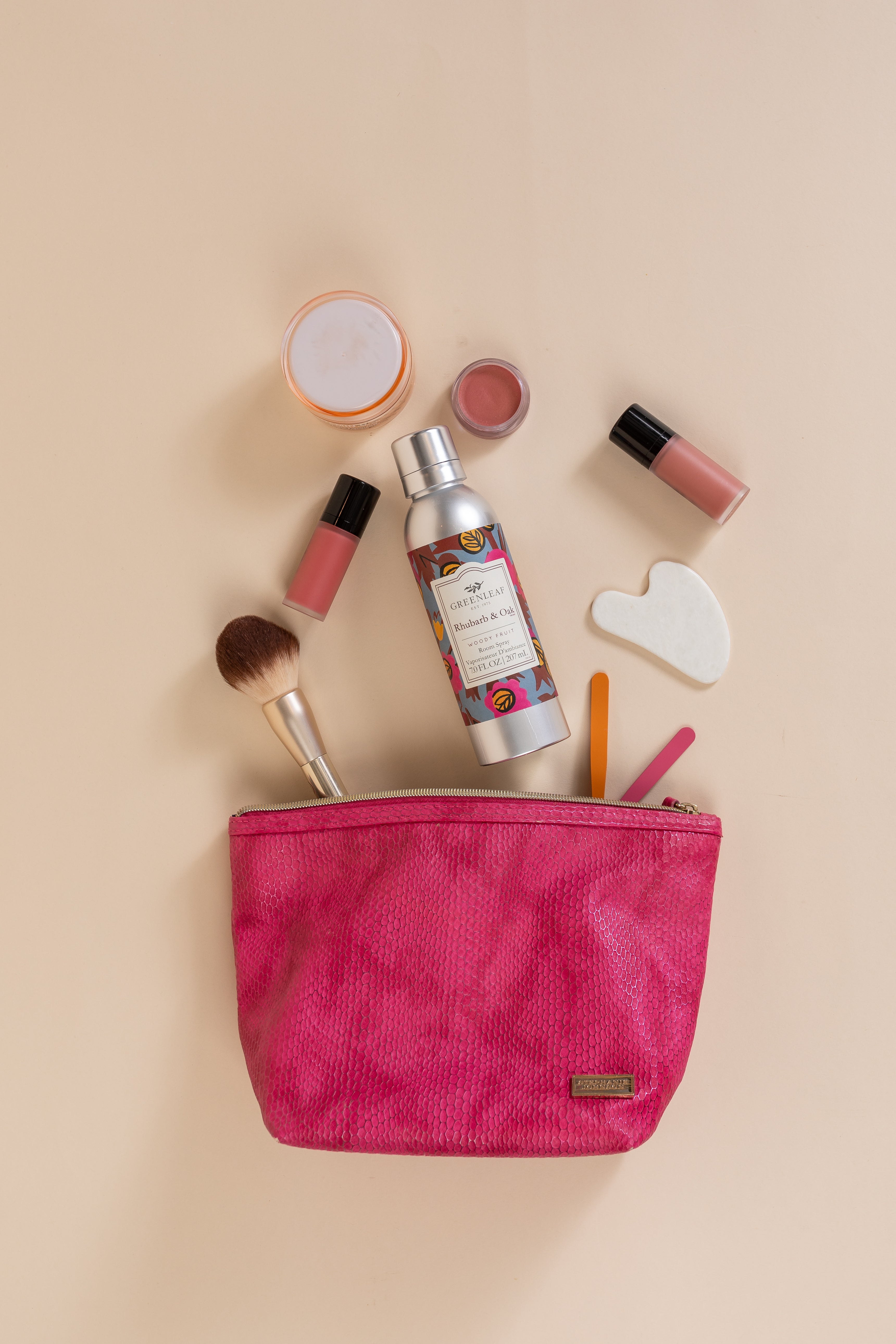 A pink makeup bag with a brush, bottle, lipsticks, small jar, white stone, nail files, and a Greenleaf Gifts Rhubarb & Oak Room Spray are neatly arranged on a beige surface.