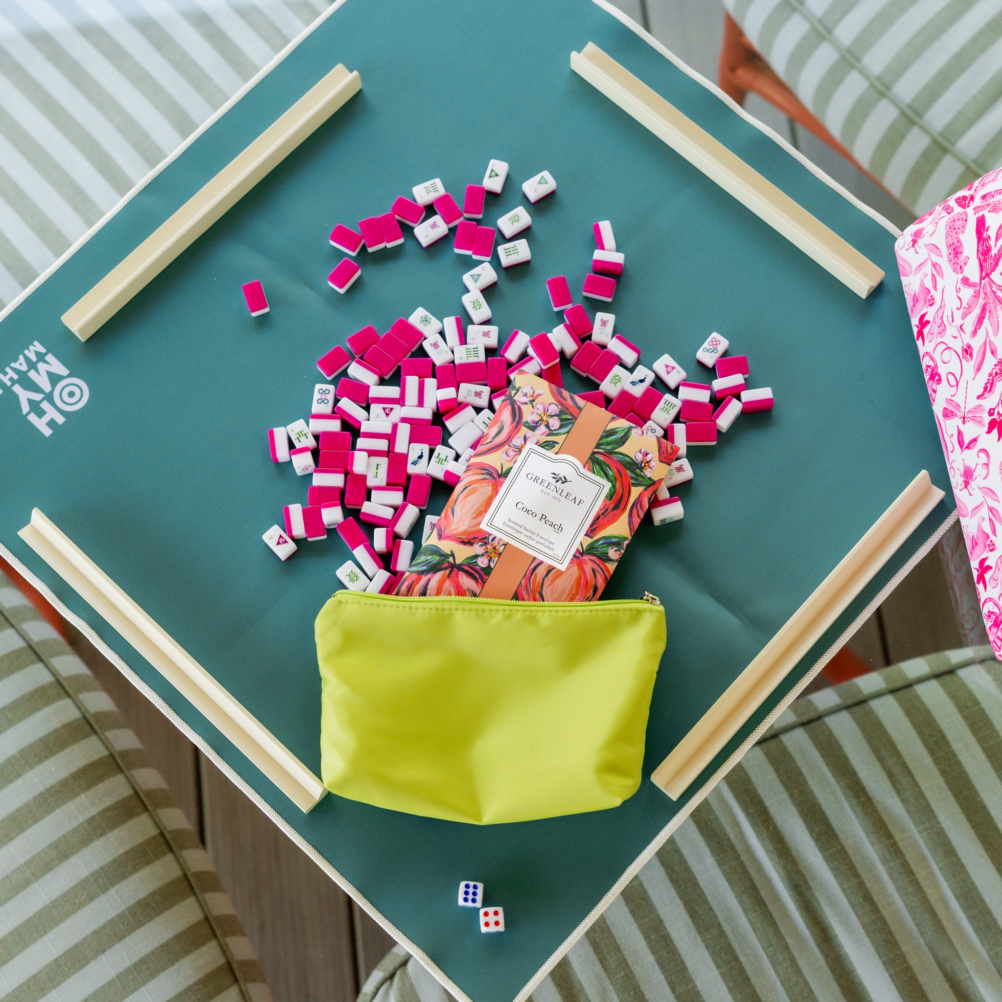 A green square table with pink and white mahjong tiles, dice, a yellow zipper pouch, and a Greenleaf Gifts Coco Peach Sachet keeps the room fresh. Four striped chairs surround the table, with tile racks set at each side.