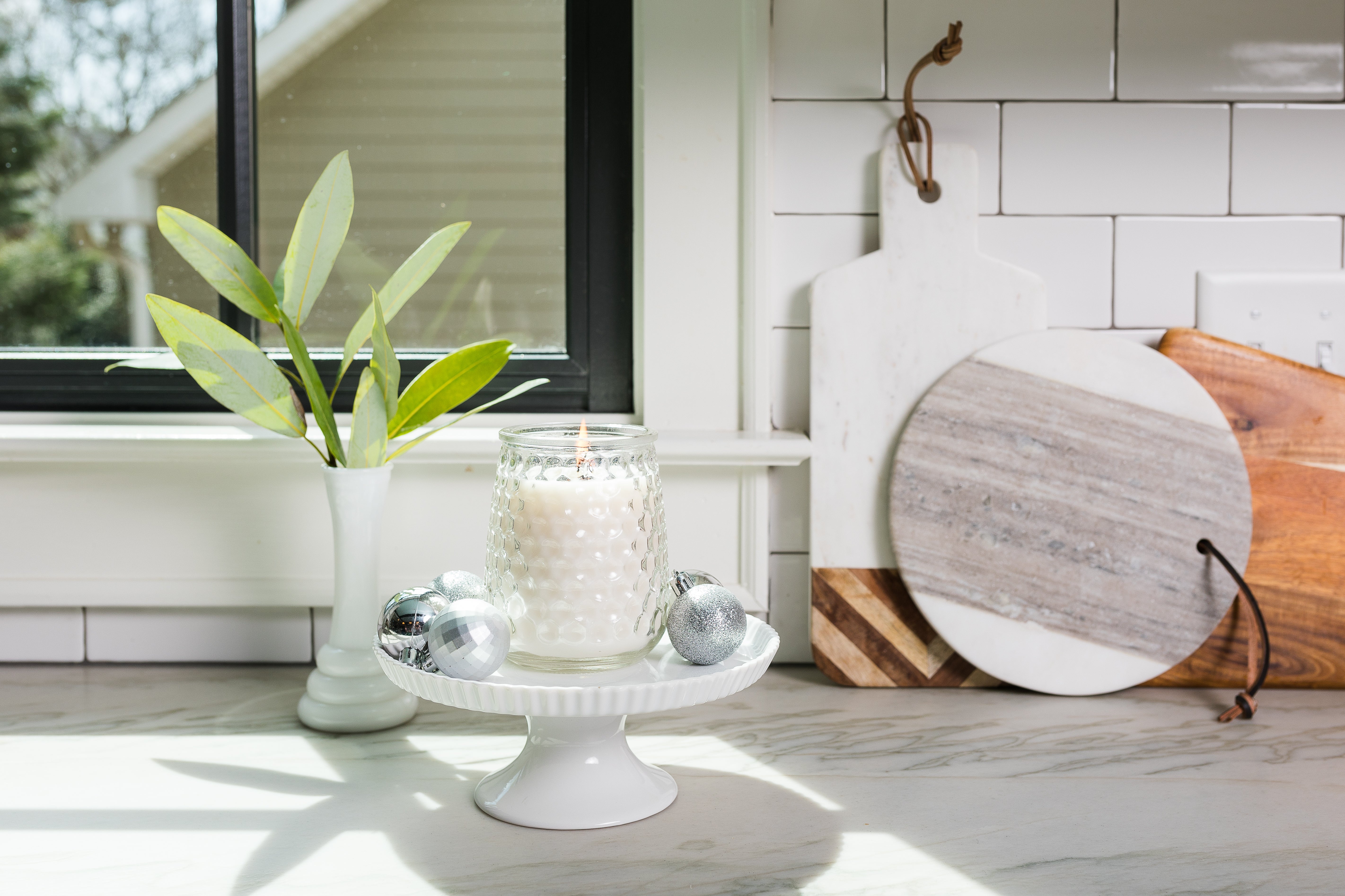 A Greenleaf Gifts Shimmering Snowberry Signature Candle in a white glass jar sits on a cake stand with three silver ornaments, with cutting boards and a vase of green leaves in the background on a sunlit kitchen counter.