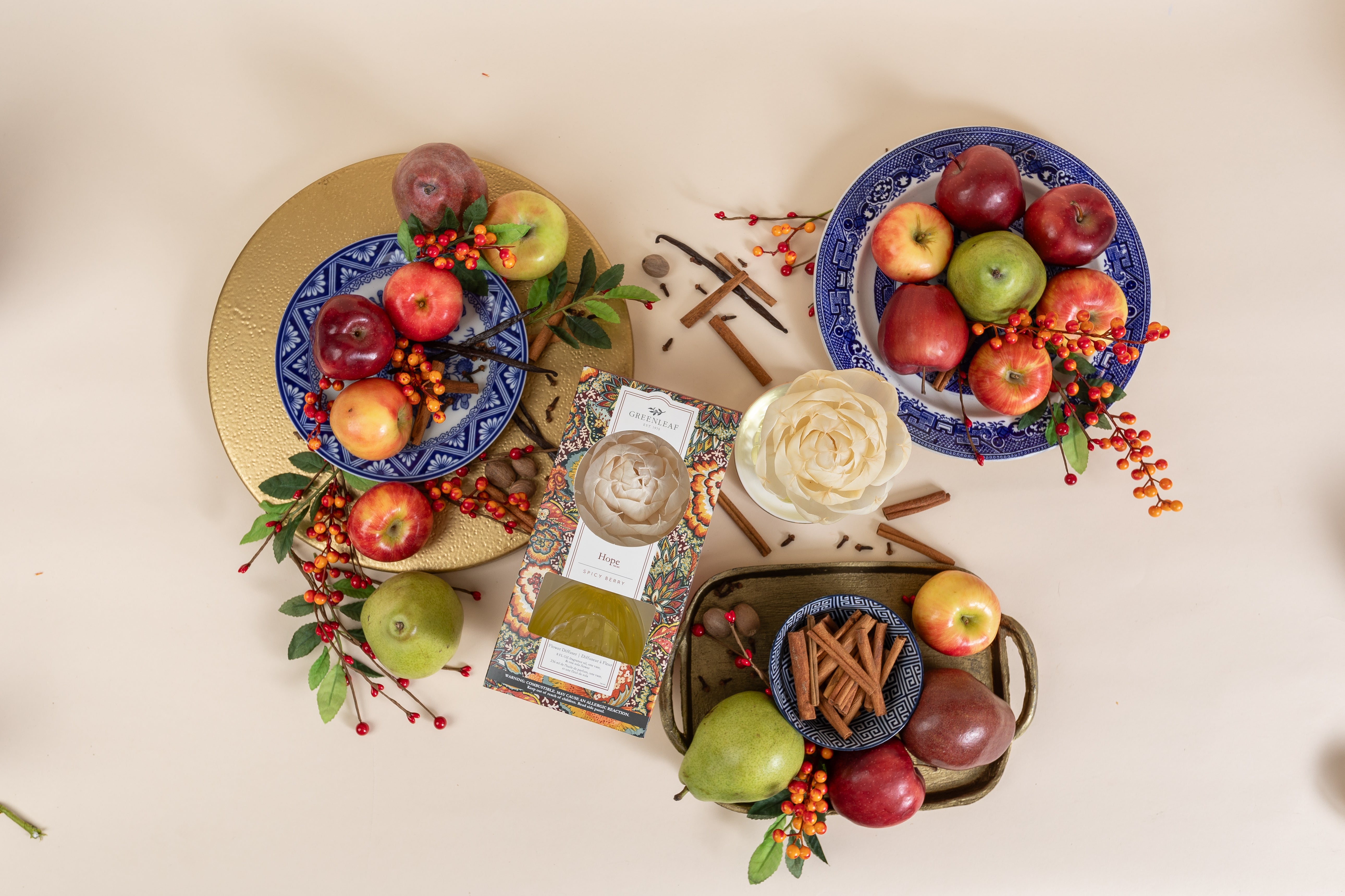 A flat lay of apples, pears, cinnamon sticks, berries, a white flower, and the Greenleaf Gifts Hope Flower Diffuser arranged on blue and gold plates and a small tray on a light surface. Product size details available for reference.