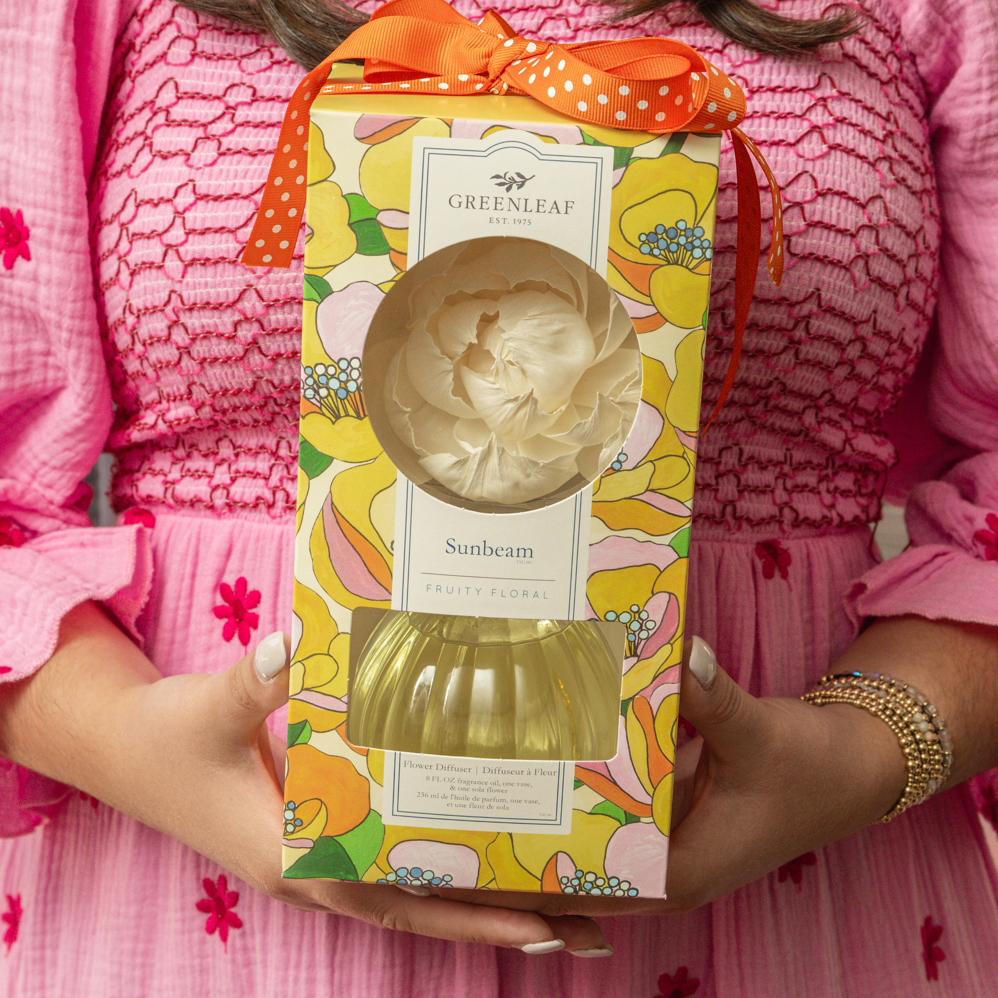A person in a pink dress holds a Greenleaf Gifts Sunbeam Flower Diffuser, which comes in colorful floral packaging with an orange ribbon and a fragrant sola paper flower displayed on top.
