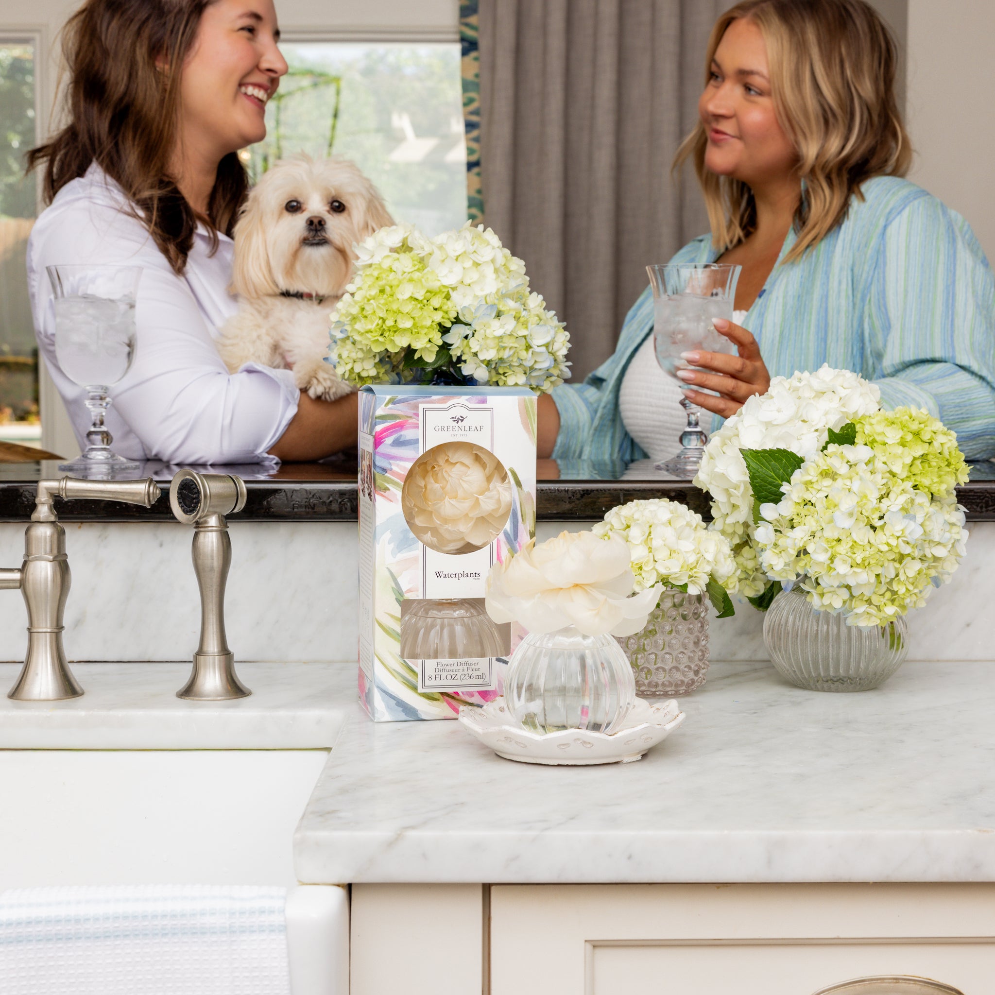 Two women smile and chat by a bathroom counter, holding glasses of water. One holds a small white dog. On the counter are green and white hydrangeas, a Greenleaf Gifts Waterplants Flower Diffuser, and a sola paper flower.
