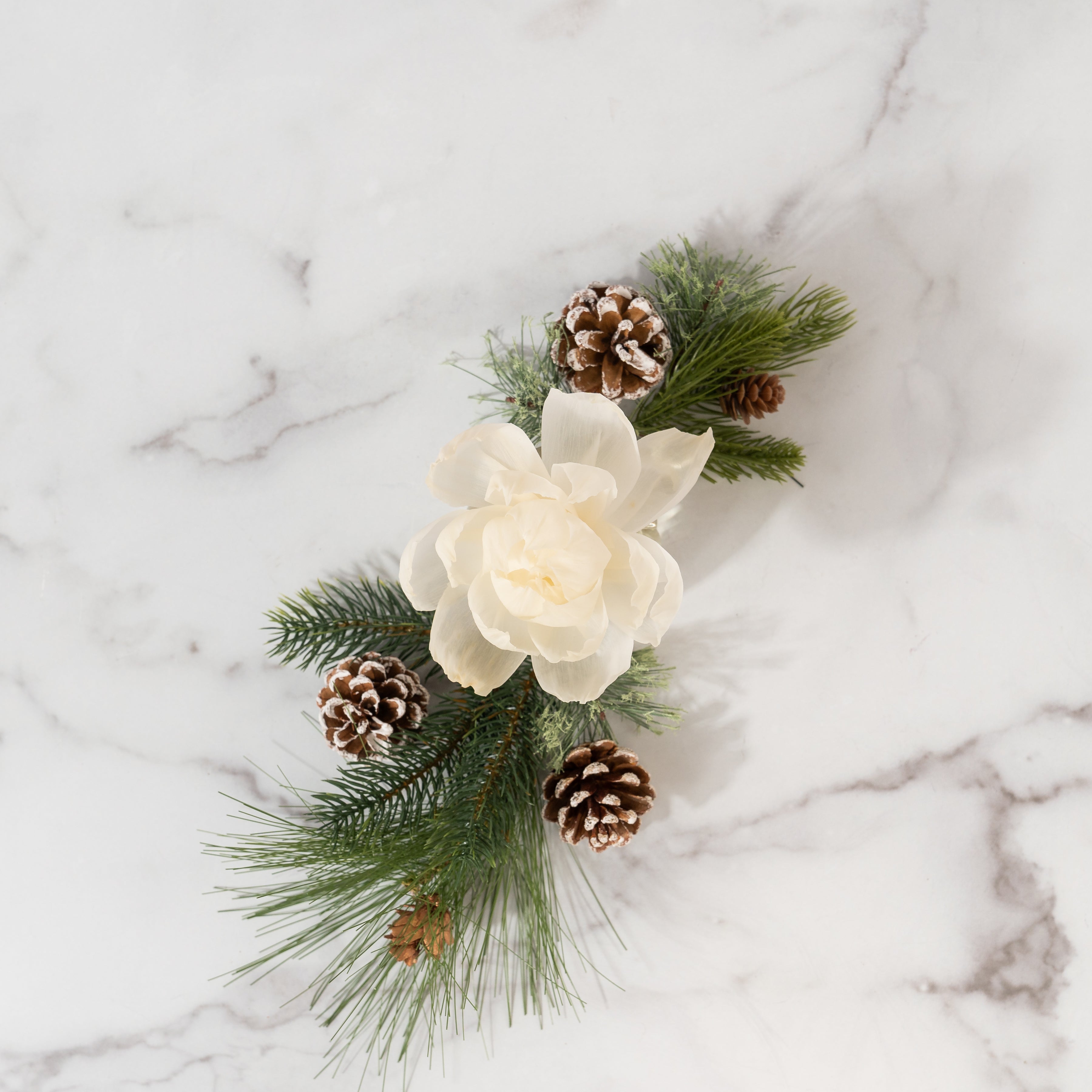 The Greenleaf Gifts Silver Spruce Flower Diffuser features a white sola paper flower with green pine branches and white-dusted pinecones, arranged on white marble—perfect for creating a charming display.