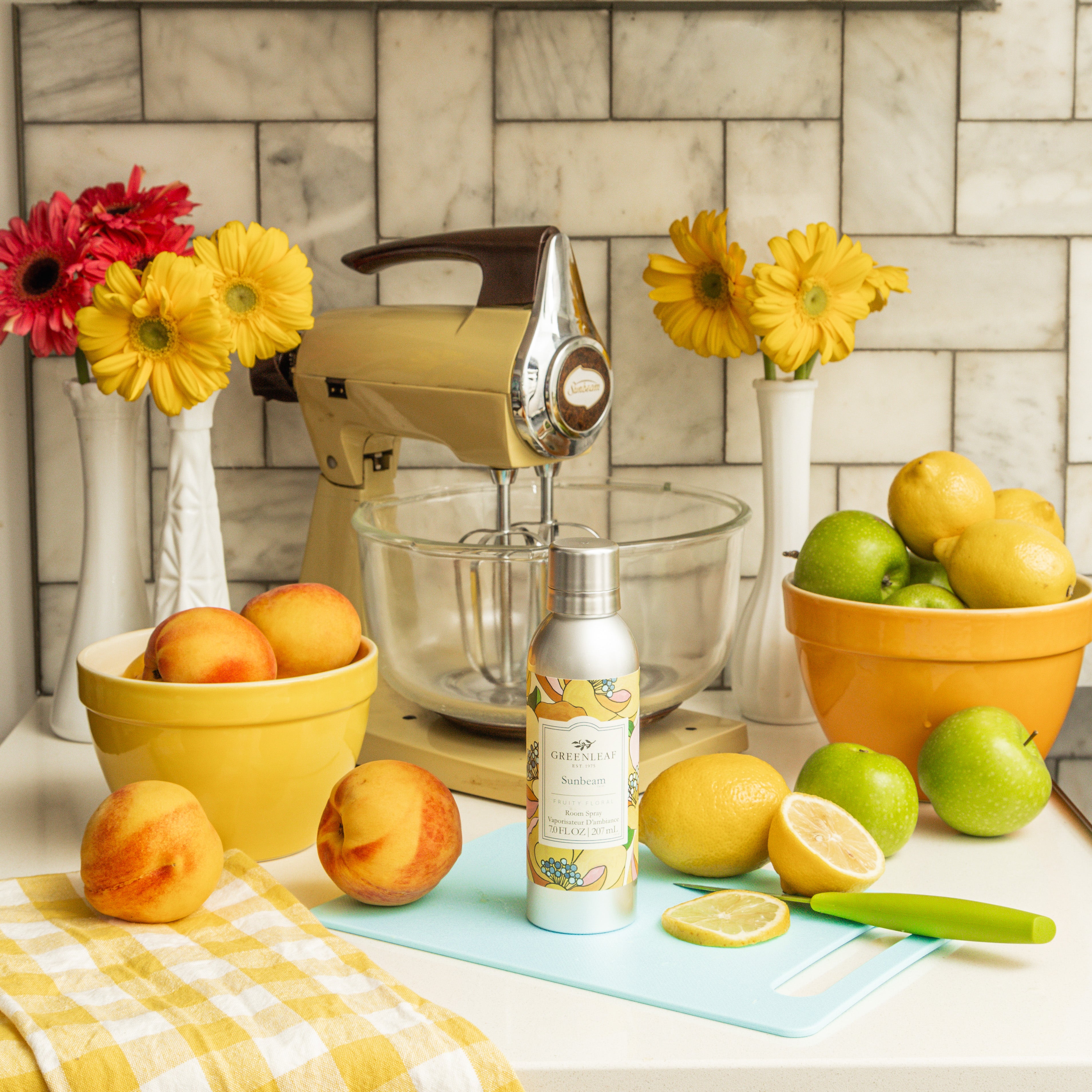 A kitchen counter with peaches, lemons, green apples, a yellow Sunbeam mixer, vases of yellow and pink flowers, a checked towel, and a Greenleaf Gifts Sunbeam Room Spray beside a cutting board with sliced lemons and a green knife.