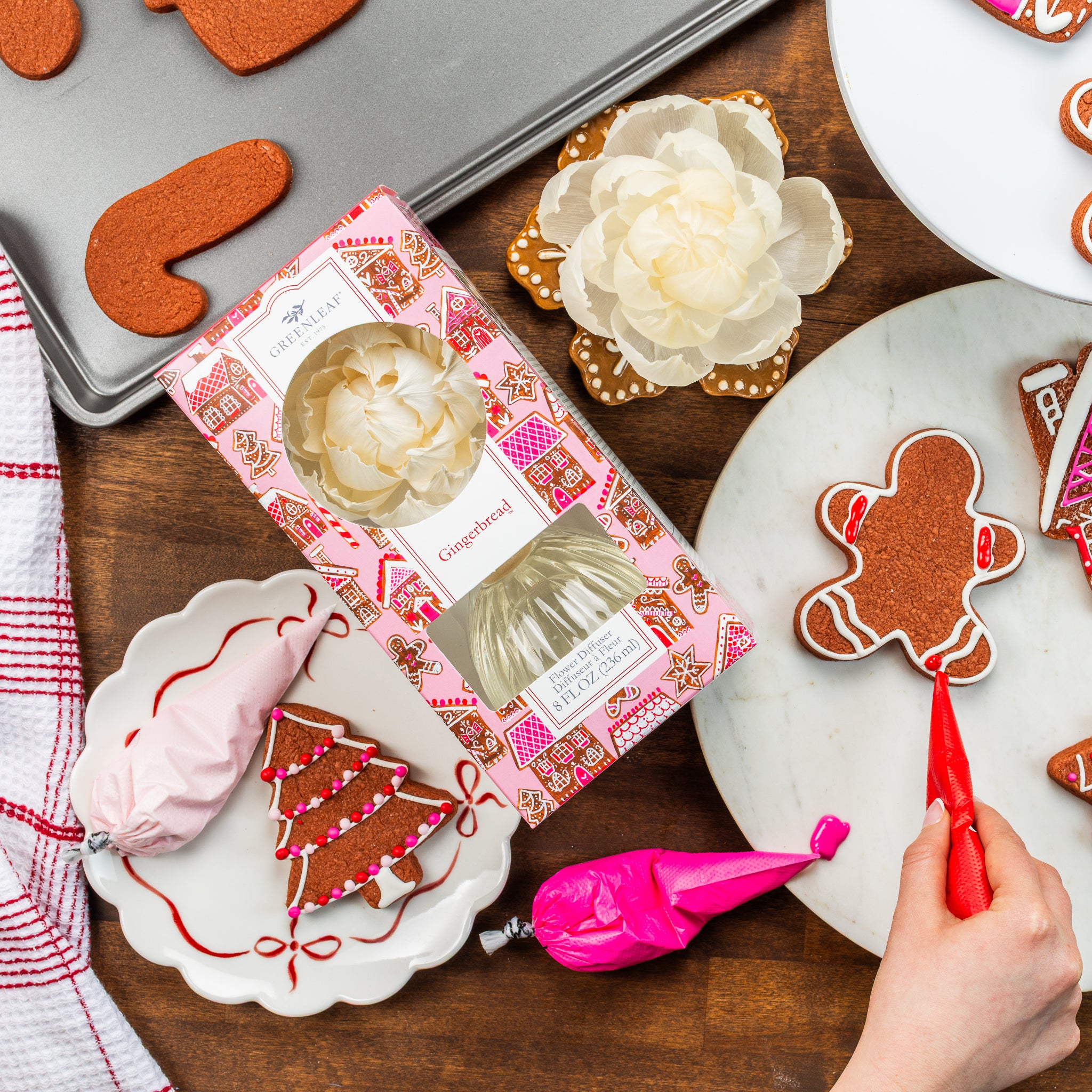 A hand decorates a gingerbread cookie with red icing. On the wooden table are a Greenleaf Gifts Gingerbread Flower Diffuser, a decorated gingerbread tree, pink and white icing bags, kitchen towels, and a cookie box.