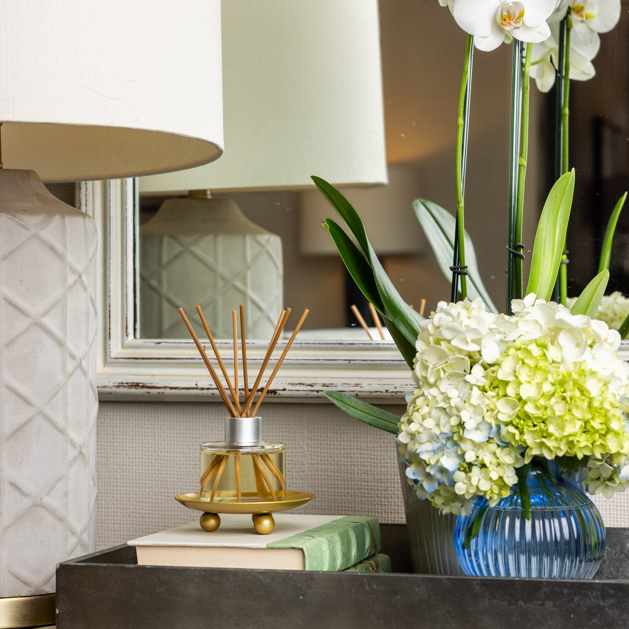 A close-up of a table with a lamp, stacked books, and a Greenleaf Gifts Waterplants Diffuser Refill Oil with ylang ylang and wooden sticks. A blue vase holds white hydrangeas and an orchid. A mirror in the background reflects part of the scene.