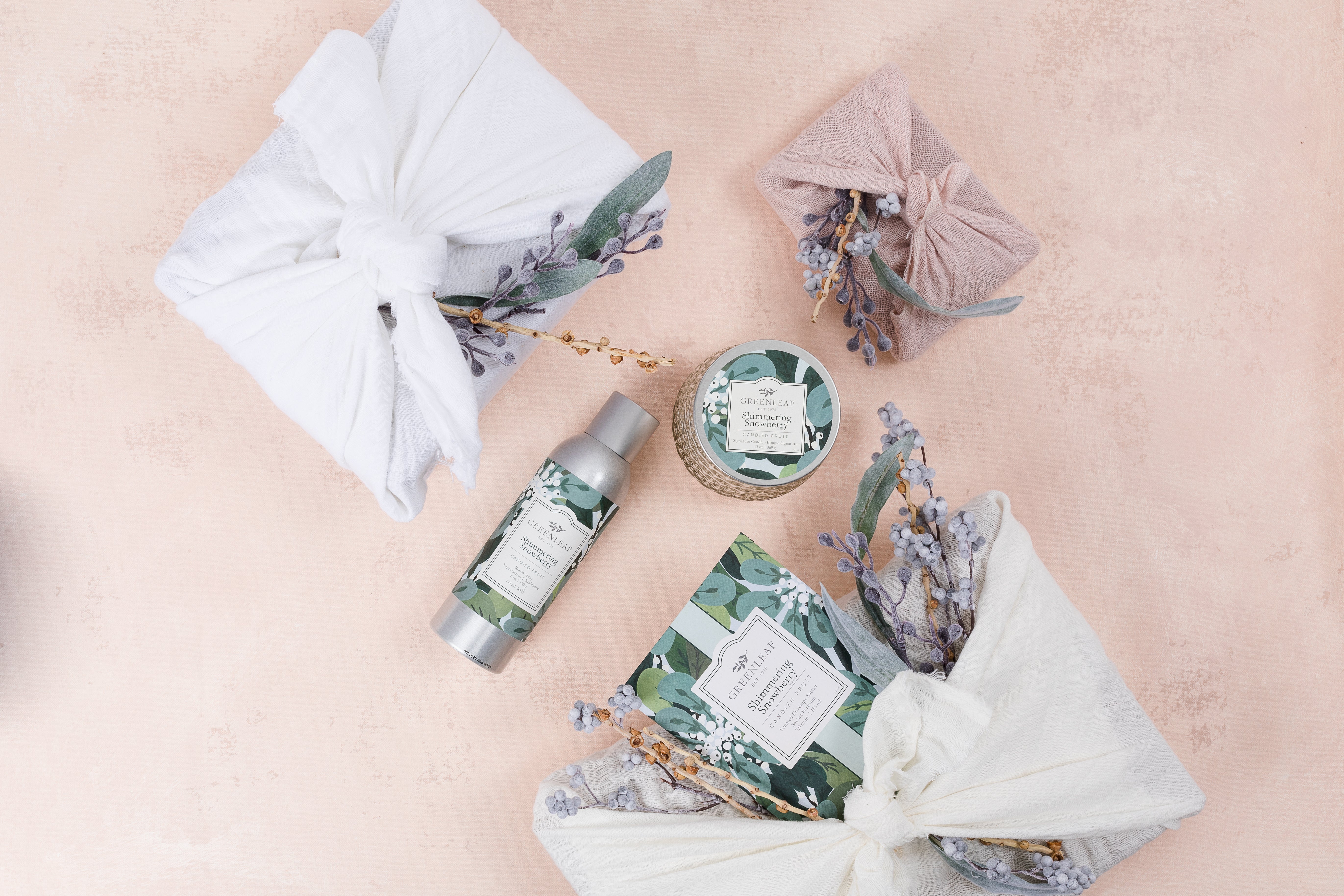 A flat lay of skincare products with botanical labels, a Greenleaf Gifts Shimmering Snowberry Signature Candle, and cloth-wrapped gifts tied with dried flowers, all artfully arranged on a soft pink background.