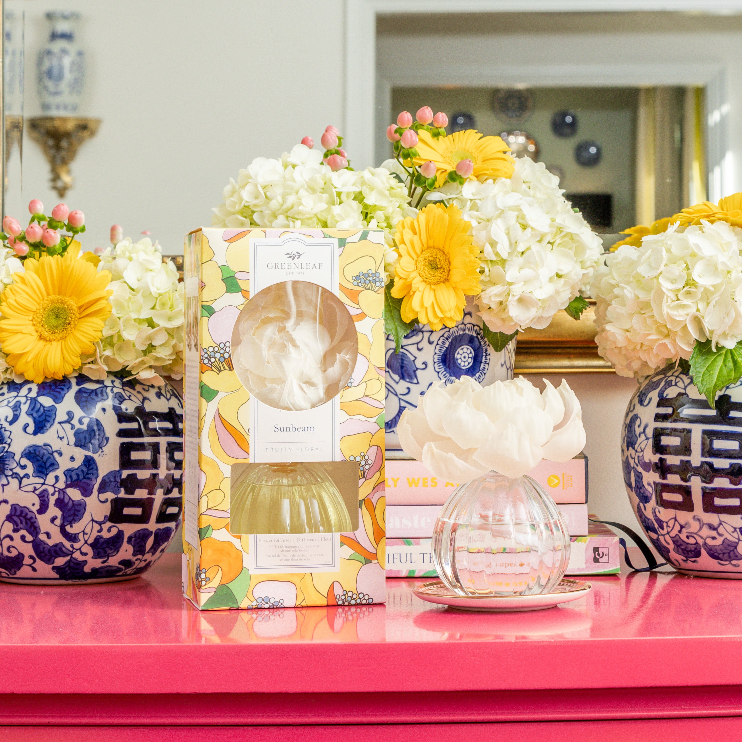 A Greenleaf Gifts Sunbeam Flower Diffuser with a sola paper flower sits on a pink table, surrounded by blue and white hydrangea vases, yellow flowers, books, and a mirror in the background.