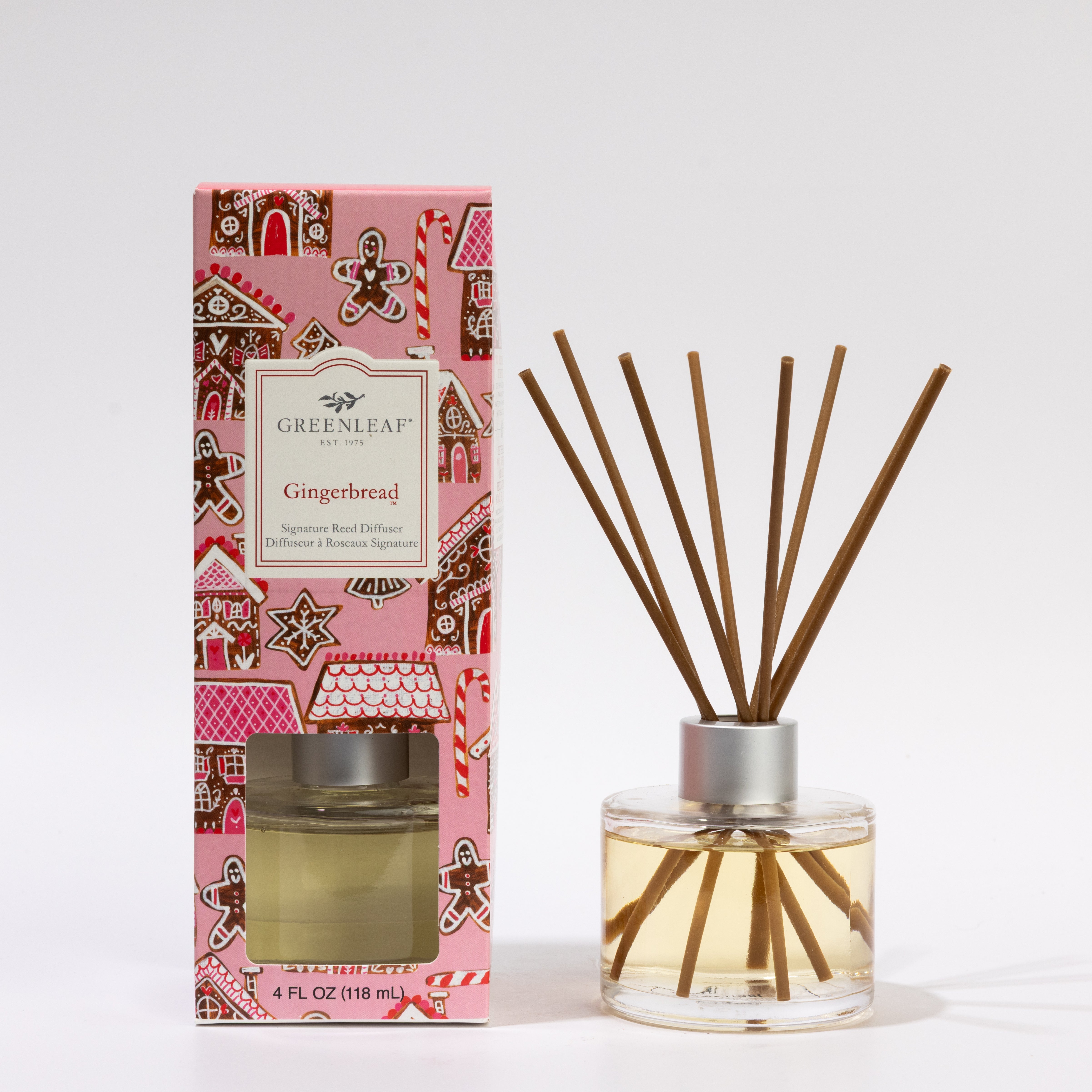 A 4 oz bottle of Greenleaf Gifts Gingerbread Reed Diffuser with several reeds, filled with light yellow liquid, sits next to its pink gingerbread-themed box. Lasts up to 30 days.