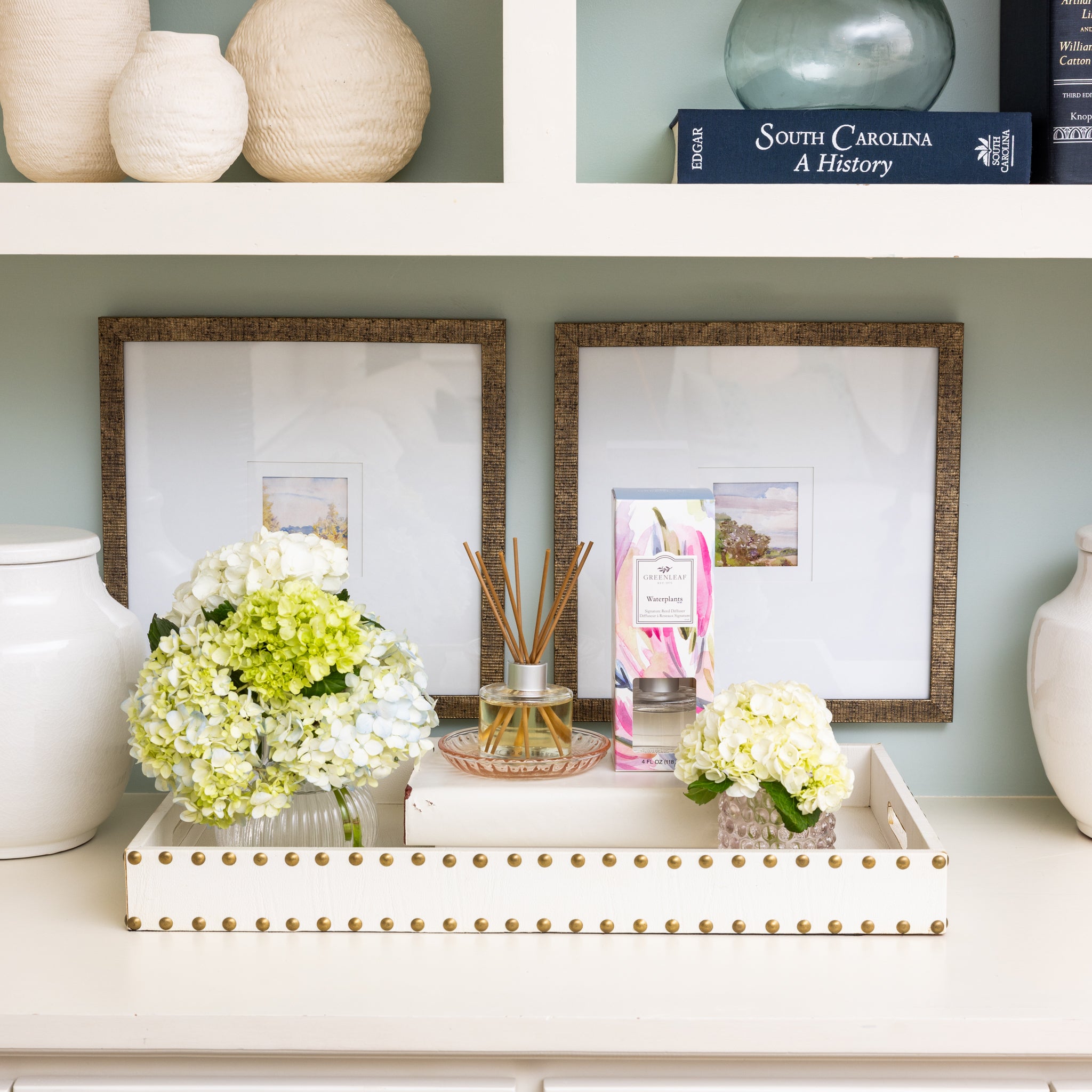 A decorative tray with flowers, a Greenleaf Gifts Waterplants Diffuser Refill Oil, and a boxed candle sits on a white shelf, flanked by white vases and framed art prints, with books and more vases displayed above.
