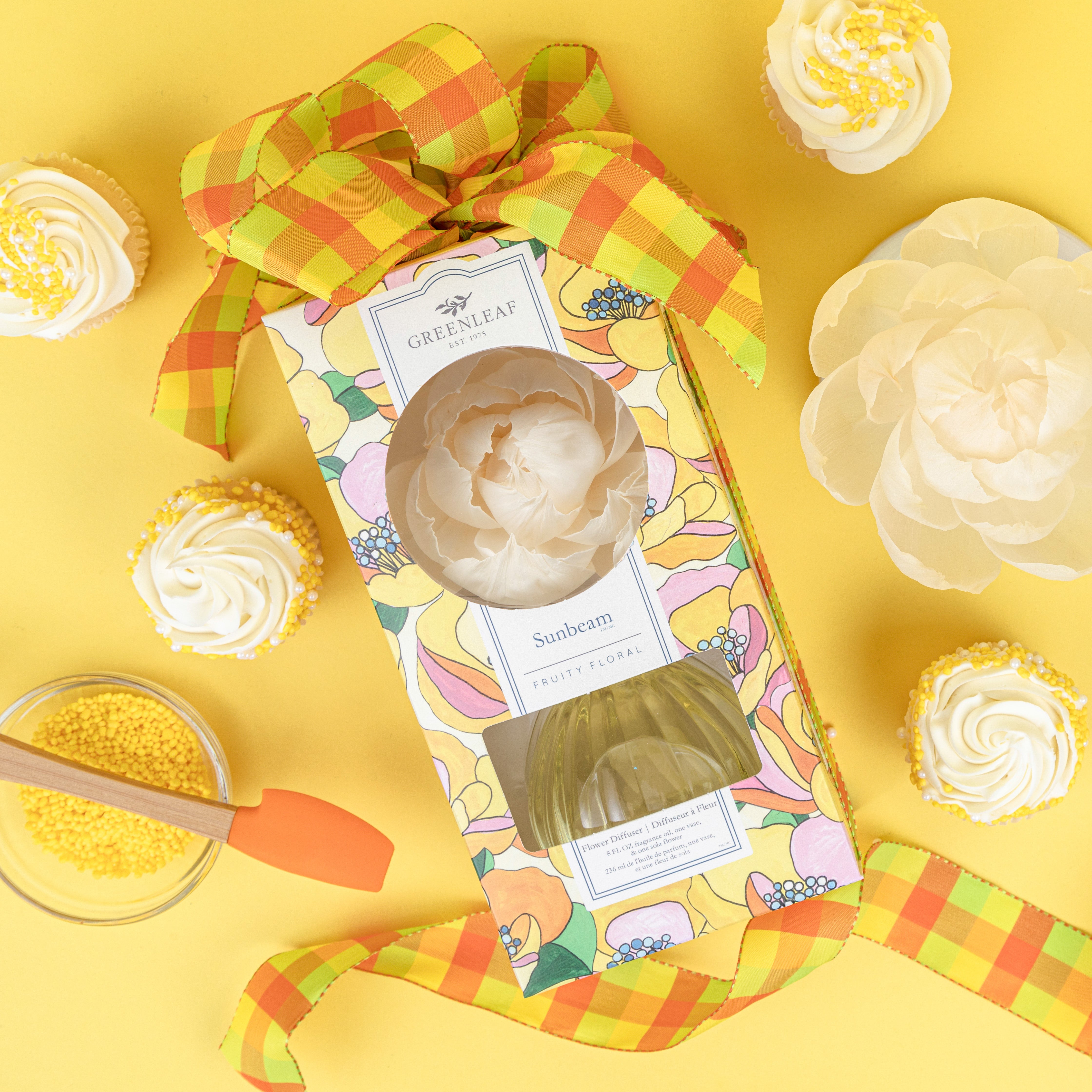 A vibrant Greenleaf Gifts Sunbeam Flower Diffuser gift box with a yellow and orange plaid ribbon sits on a yellow background, surrounded by white cupcakes with yellow sprinkles, a glass bowl of sprinkles, spatula, and sola paper flower.