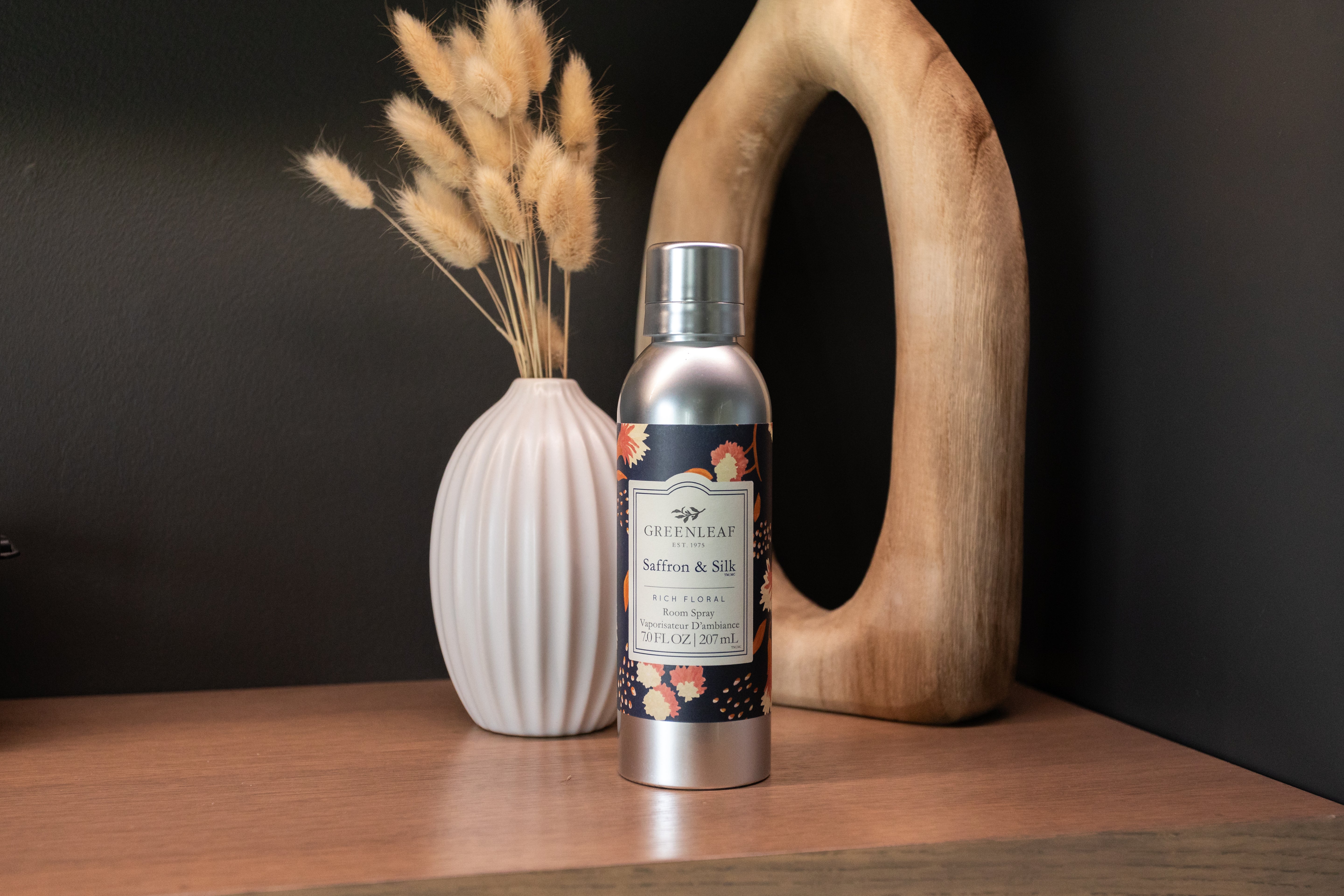 A silver can of Greenleaf Gifts Saffron & Silk Room Spray sits on a wooden surface beside a white vase with dried grass and a decorative wooden ring, set against a dark background.