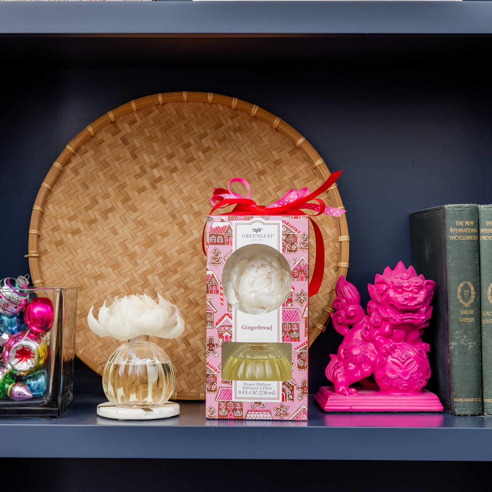 A shelf displays Greenleaf Gifts' Gingerbread Flower Diffuser, featuring a sola paper flower, plus colorful ornaments, a bright pink decorative lion, and a green book.