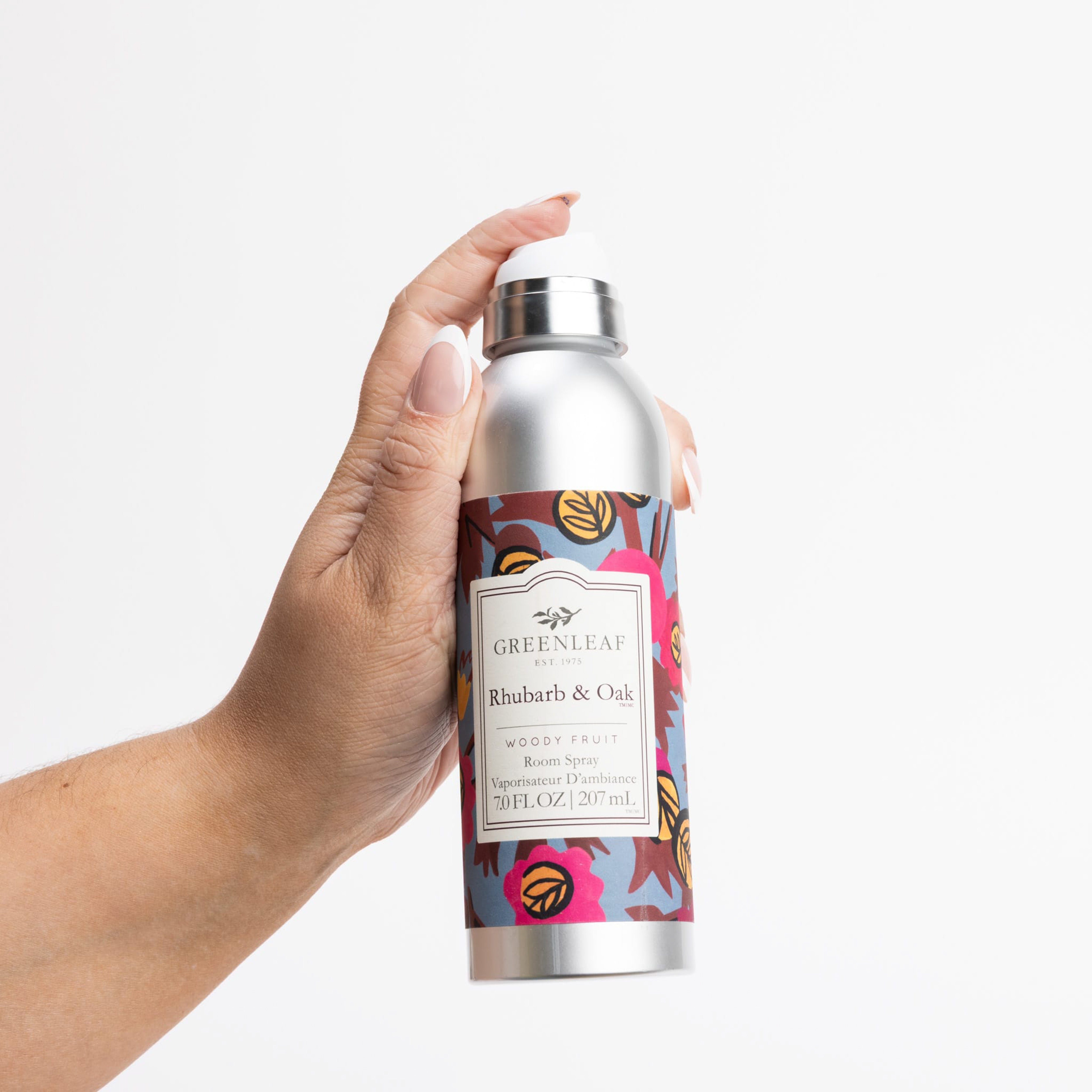 A hand holds a silver non-aerosol Greenleaf Gifts Rhubarb & Oak Room Spray with a vibrant leafy label design, set against a plain white background.