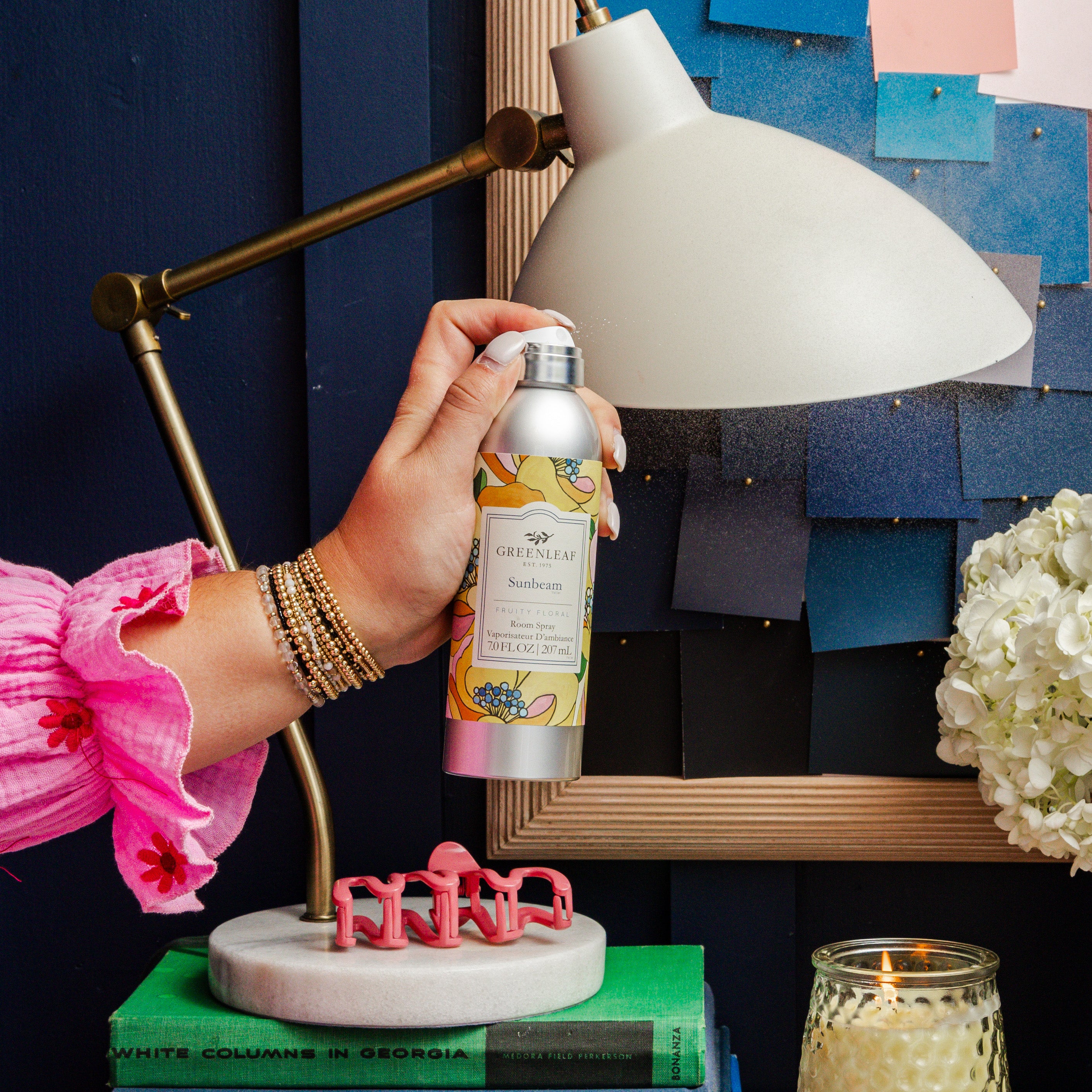 A person in a pink blouse and gold bracelets holds a Greenleaf Gifts Sunbeam Room Spray beside a modern desk lamp, books, pink glasses, and a lit candle on a white marble stand against a dark blue background.
