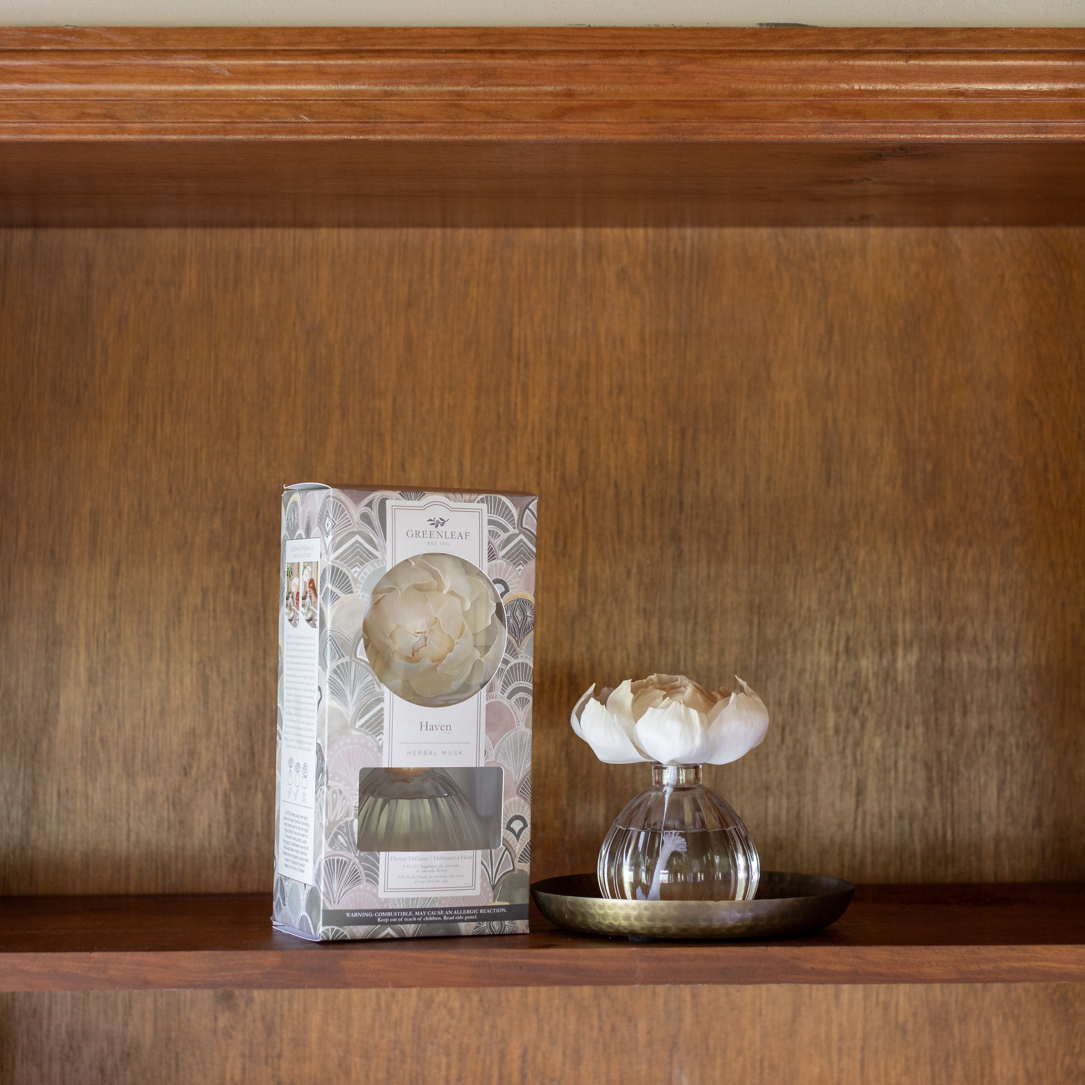 The Haven Flower Diffuser by Greenleaf Gifts, featuring a white sola paper flower on a round tray, sits beside its patterned box on a wooden shelf. The simple wooden background highlights its elegant design and delicate fragrance.