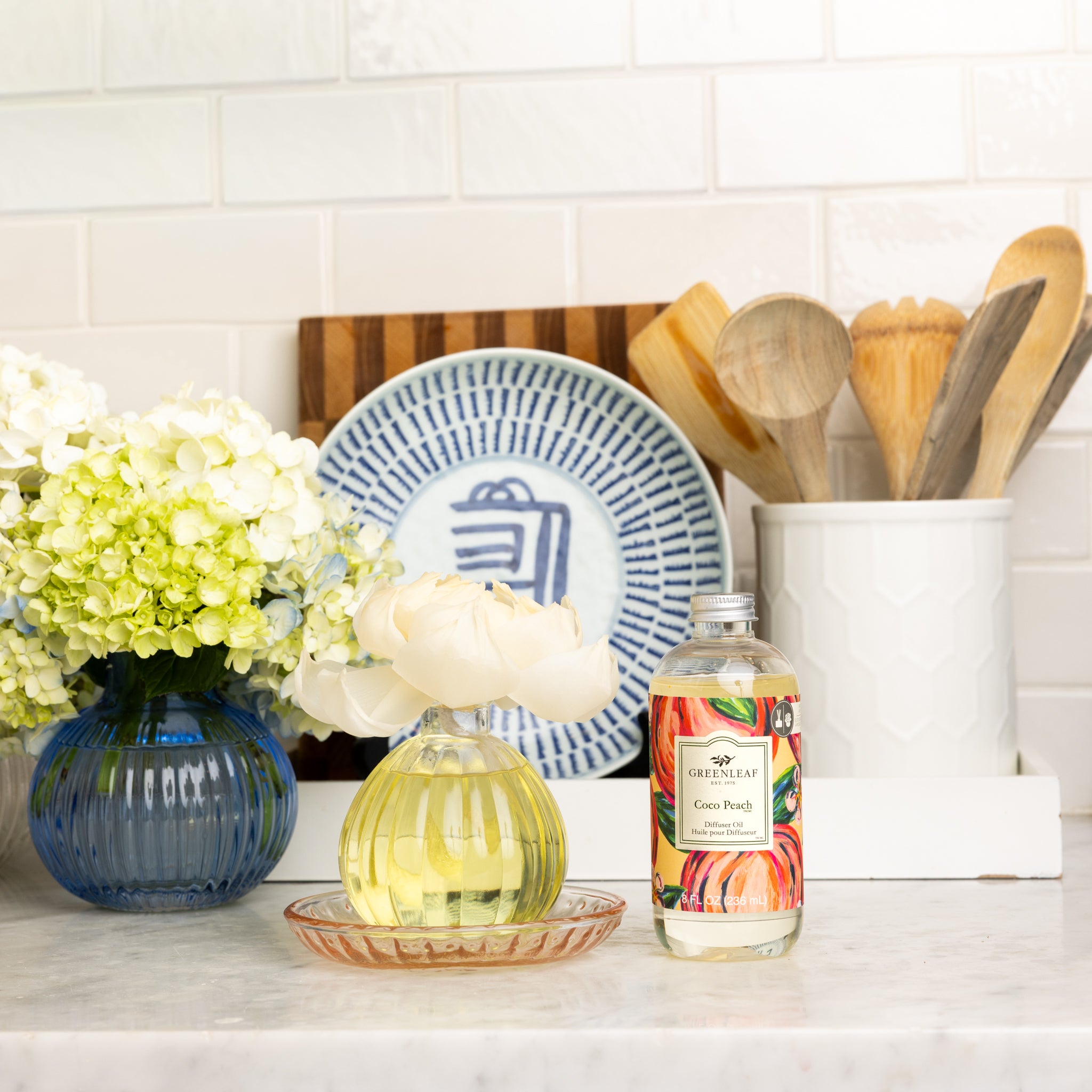 A kitchen counter with a vase of white flowers, a Greenleaf Gifts Flower Diffuser using Coco Peach Diffuser Oil, a blue-patterned plate, and a white utensil holder filled with wooden spoons.