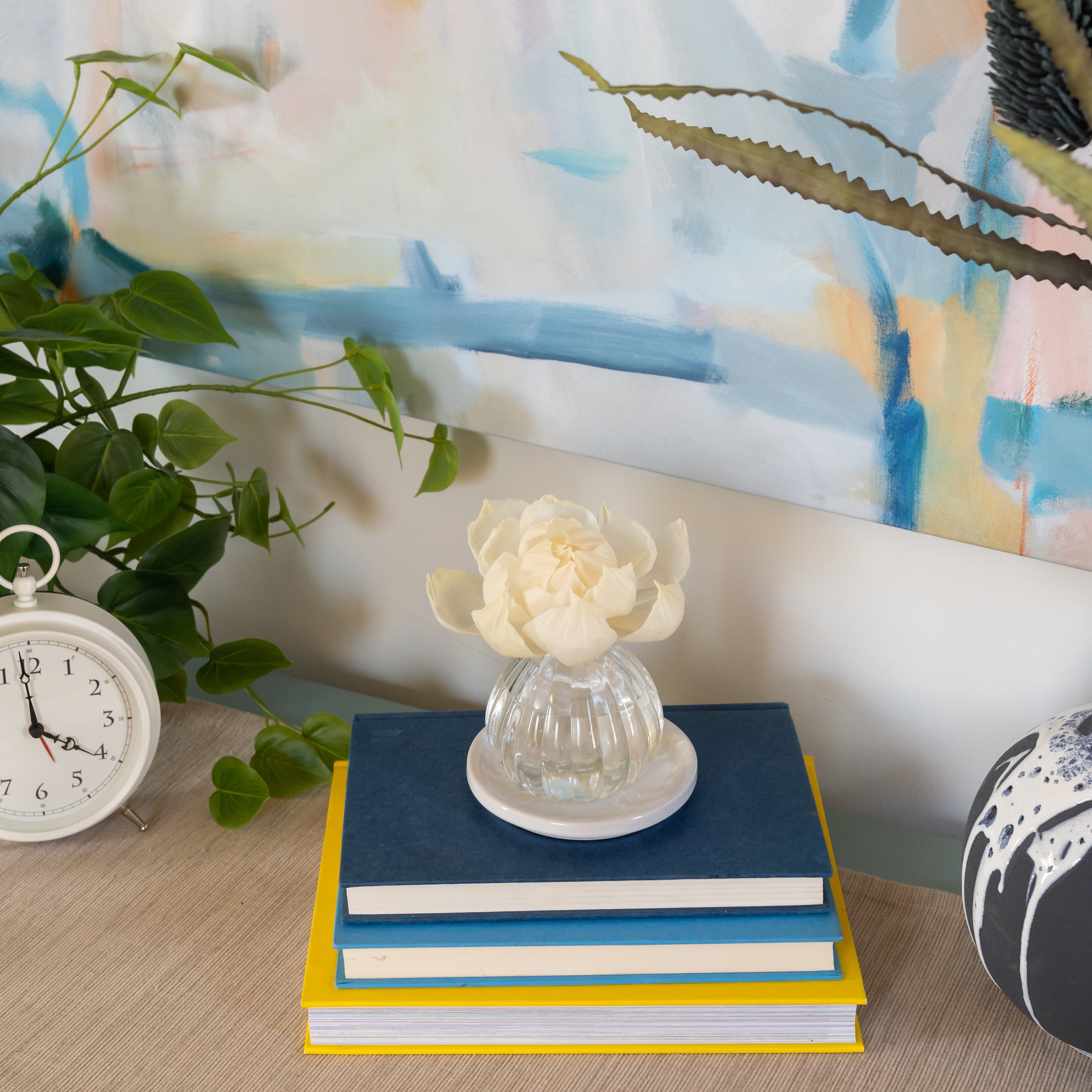 The Flower Diffuser-Seaspray by Greenleaf Gifts features white blooms in an 8 oz glass vase, styled with blue and yellow books, a vintage alarm clock, leafy plant, and abstract art. Enhances any space for up to 60 days.